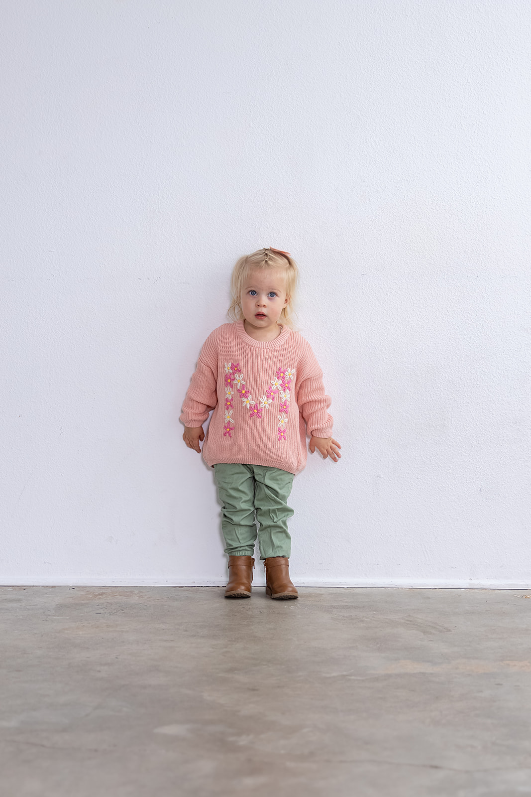 Young girl with blonde hair in a pink sweater, green pants, and brown boots standing against a plain white wall.
