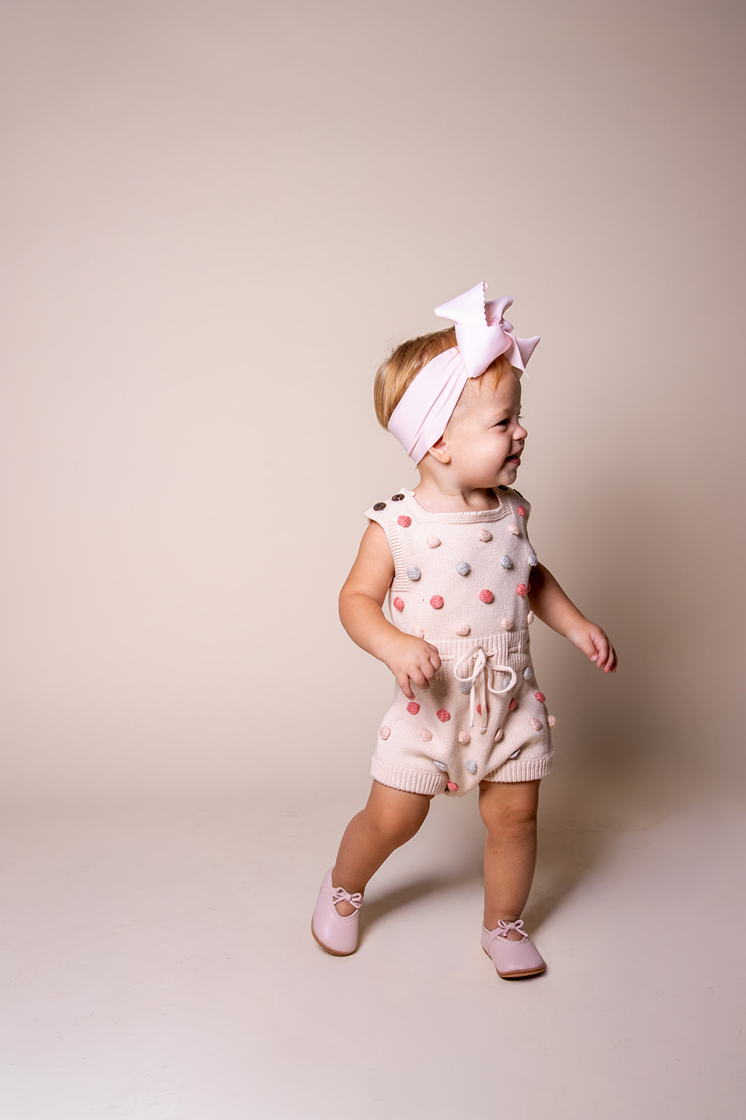 Young girl with a large bow headband, wearing a polka dot romper, standing and smiling, against a plain background.