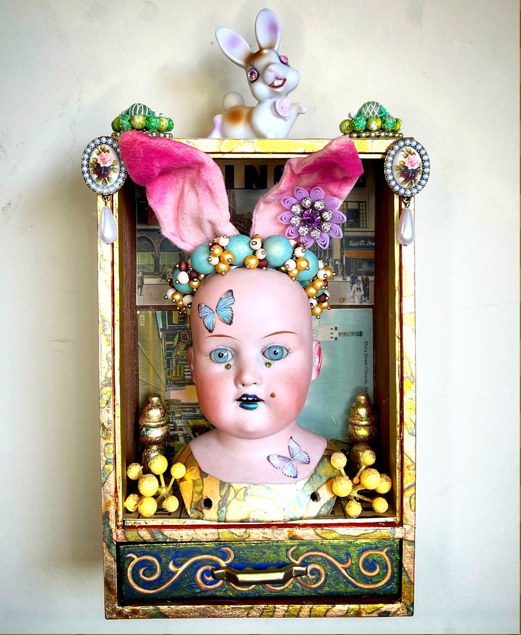 Decorative display with a doll's head, large pink bow, butterfly accessories, and a rabbit figurine on top, set in a wooden frame.
