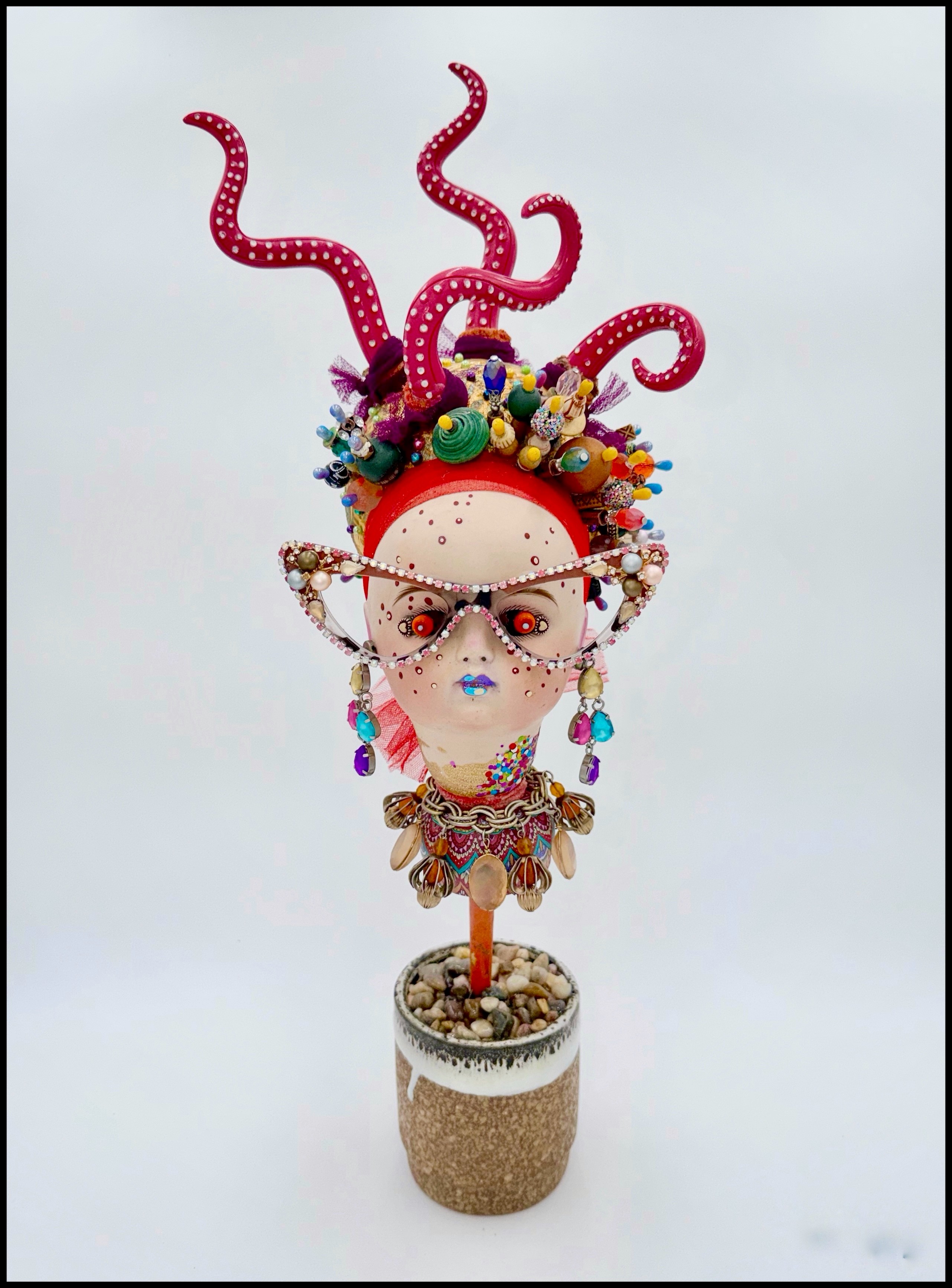 Decorative bust with colorful headpiece, glasses, and jewelry in a pot, against a plain background.