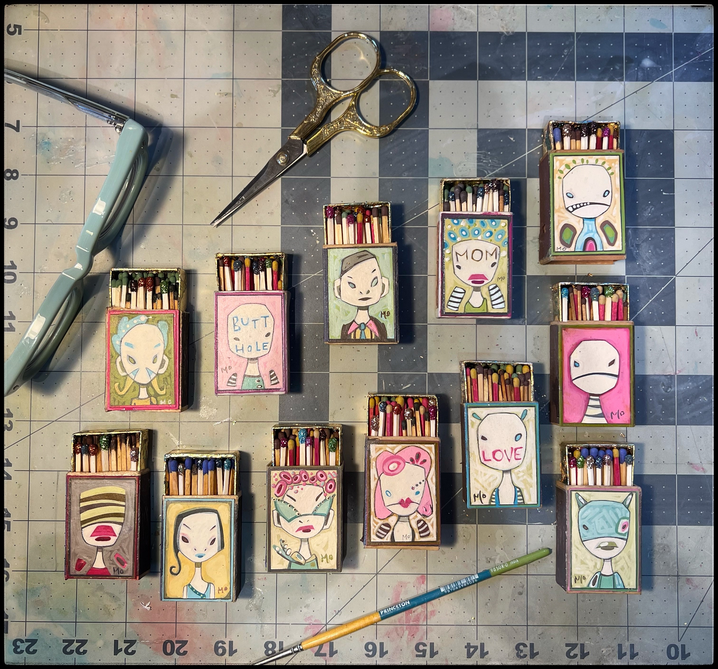 Small boxes with hand-drawn faces and messages, surrounded by scissors, ruler, and a paintbrush on a tiled surface.