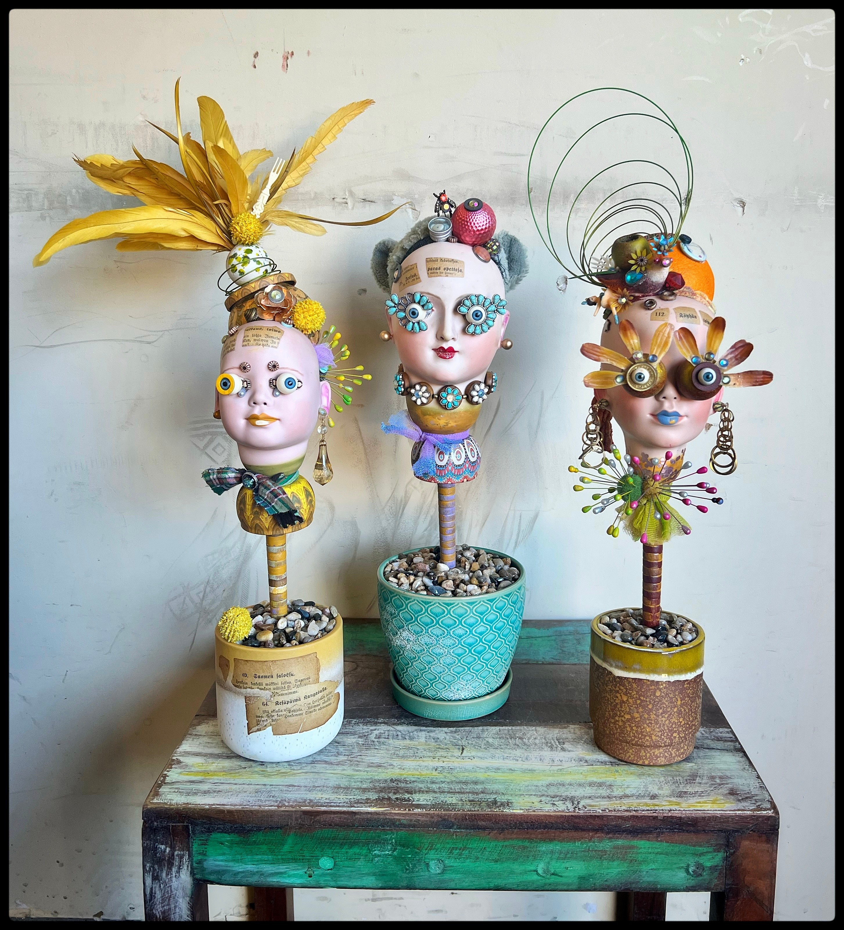 Three decorative figurines with colorful accessories and exaggerated features on a wooden table against a white wall.