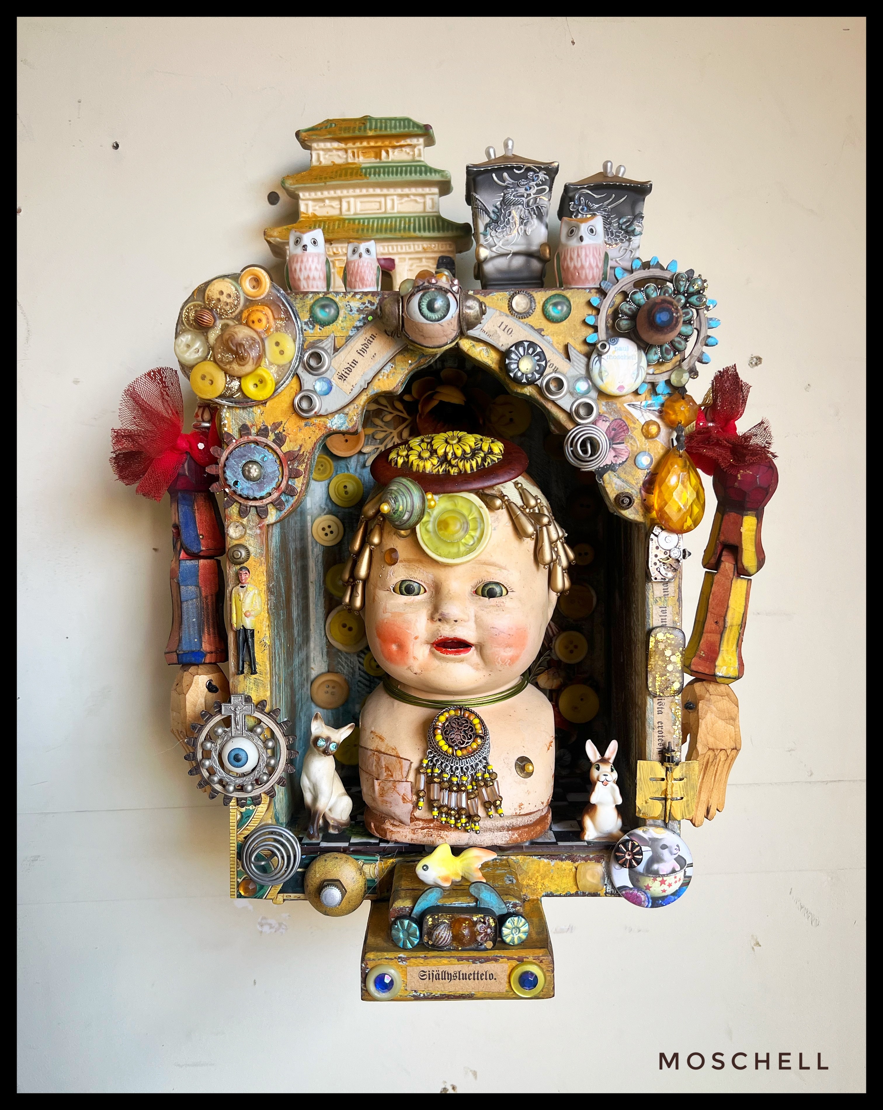 Colorful sculpture of a baby with decorative elements, books, and objects around it, mounted on a white wall.