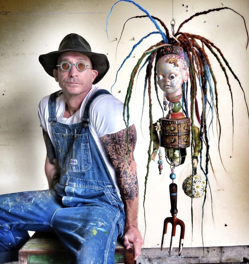 Man with glasses, hat, and tattoos sitting next to a colorful, artistic sculpture with long, multicolored dreadlocks and a face mask.