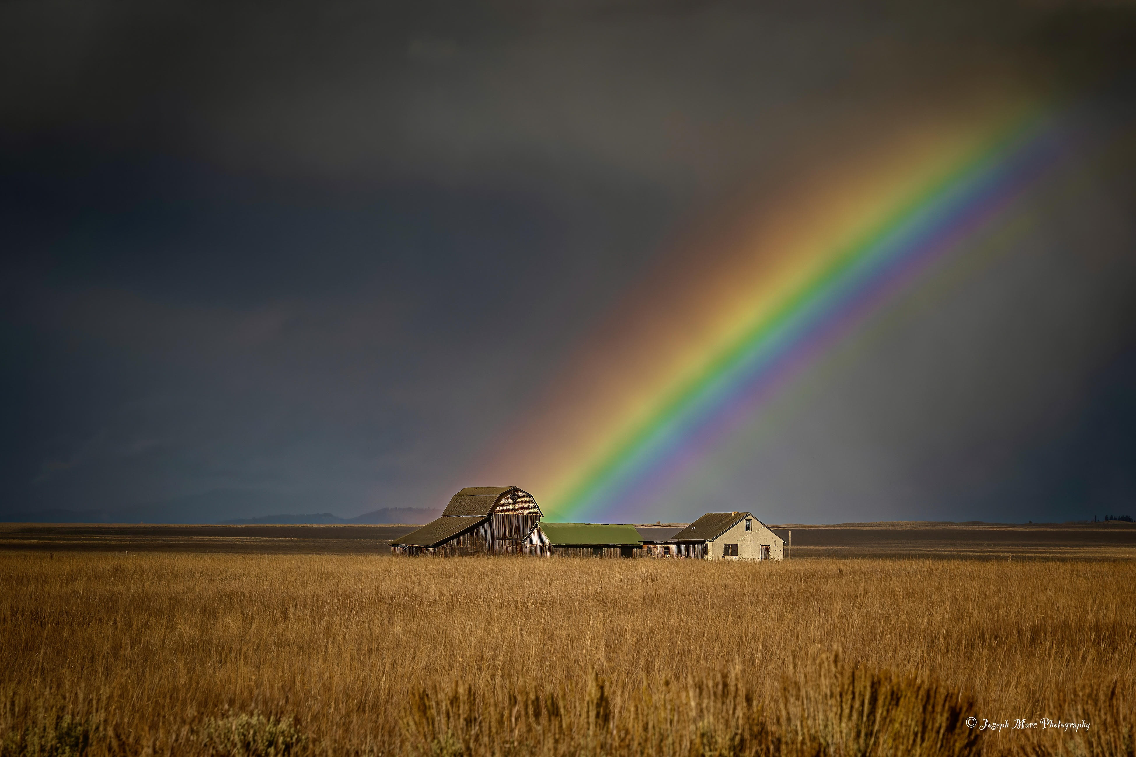 A rainbow arches over farm buildings in a field with dark clouds in the sky.