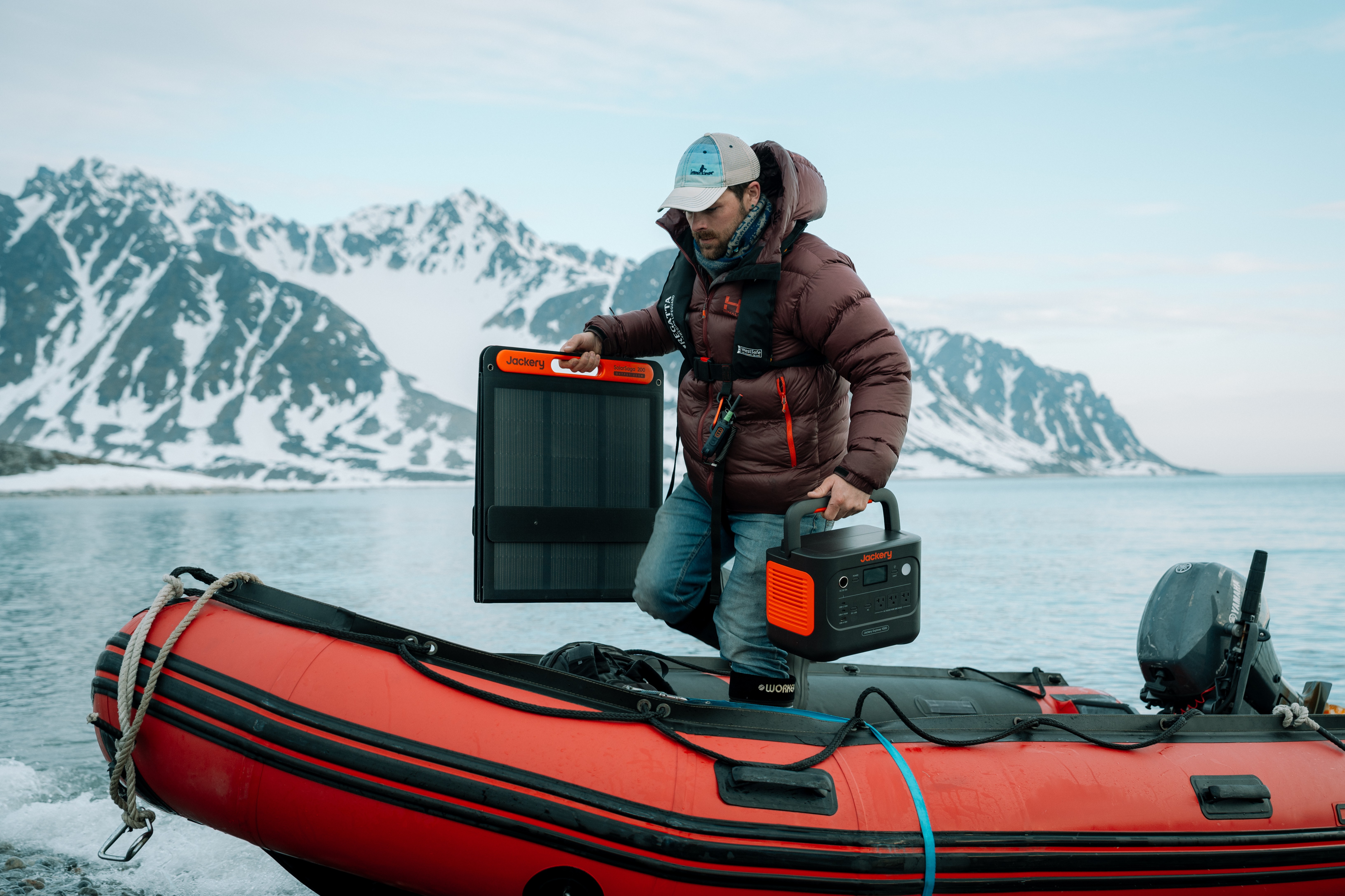 Person in winter clothing on a boat with equipment, snowy mountains in the background, water, and sky.