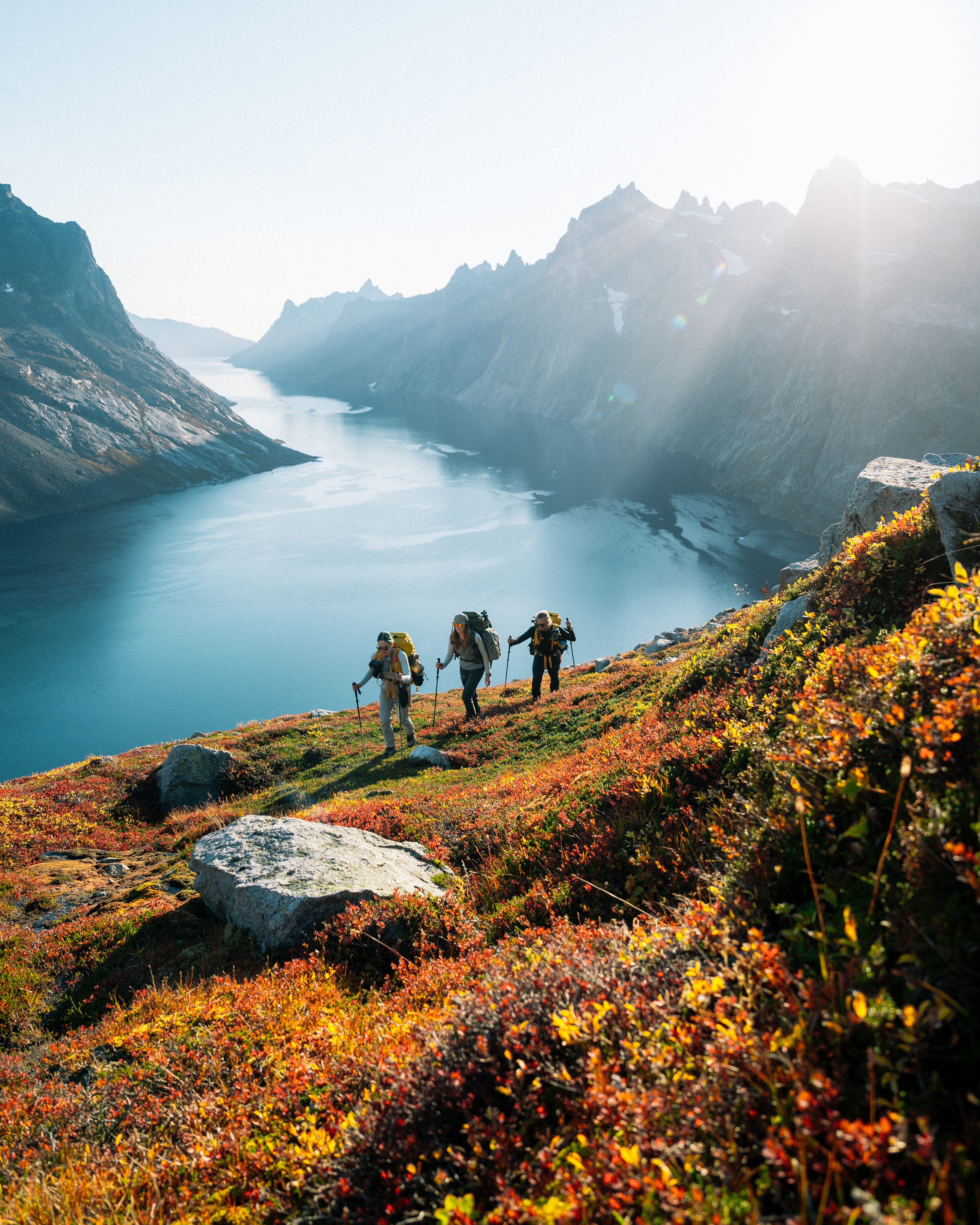 Four hikers walking along a colorful hillside overlooking a river in a mountainous landscape, with sunlight shining from the right.