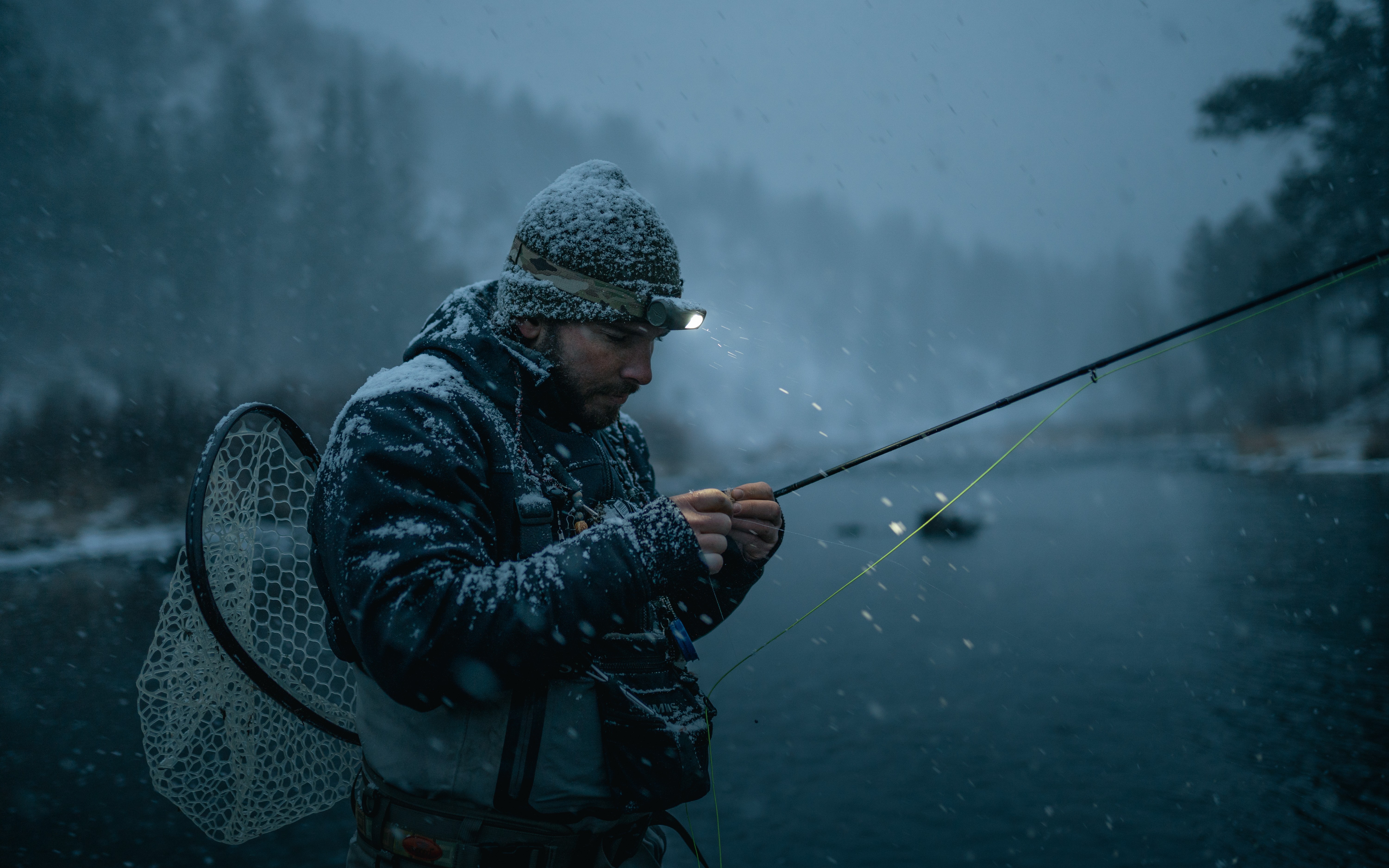 Person fishing in a river during rainy weather, wearing a hat and waterproof clothing, with a backpack and fishing rod.