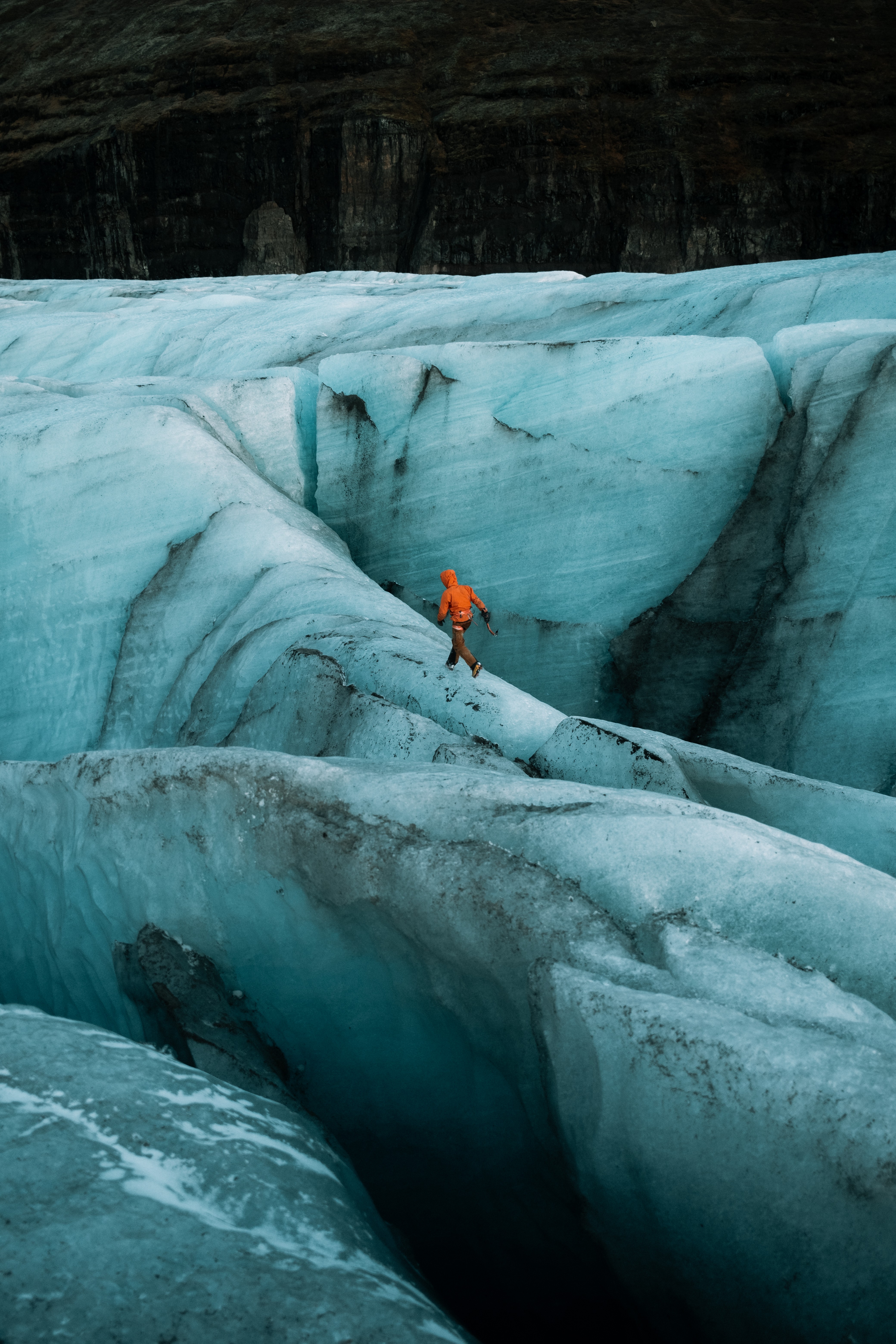 Person in orange jacket walking on icy glacier with deep crevasses and dark rocky background.