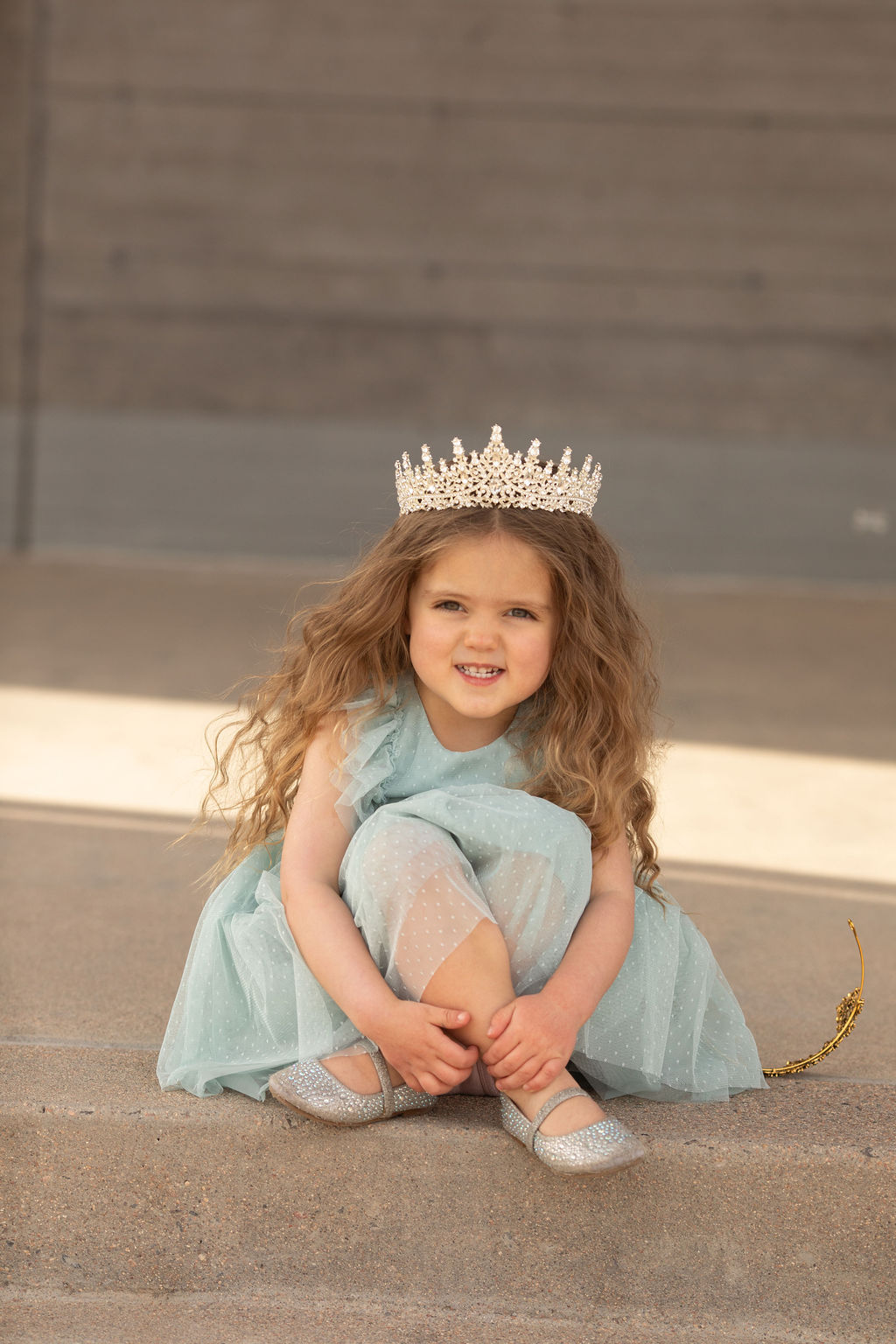 Young girl with curly hair wearing a crown and a light blue dress, sitting on a step smiling.