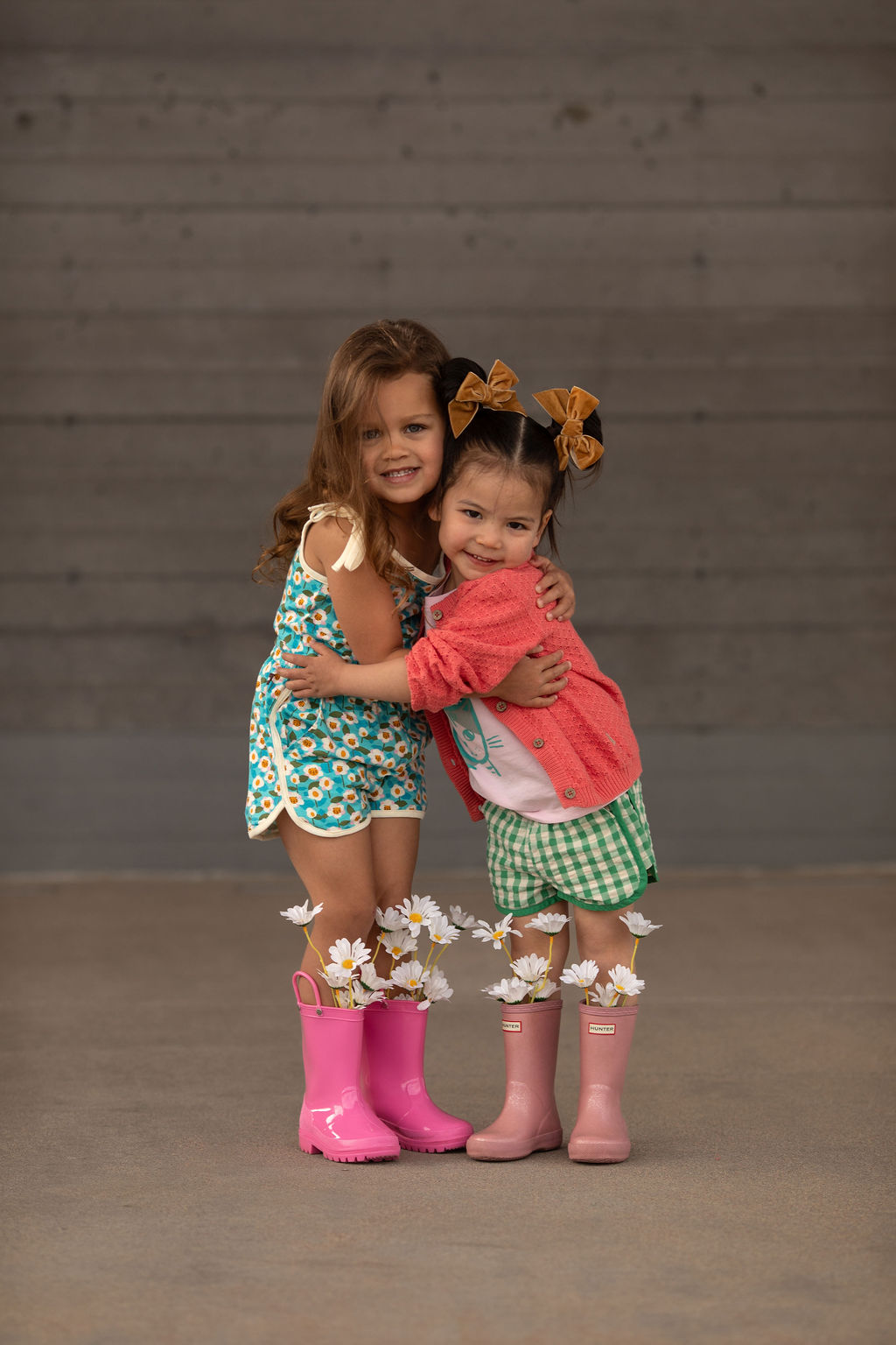 Two young girls hugging, smiling, wearing colorful dresses and pink rain boots, standing on a plain floor with a gray wall background.
