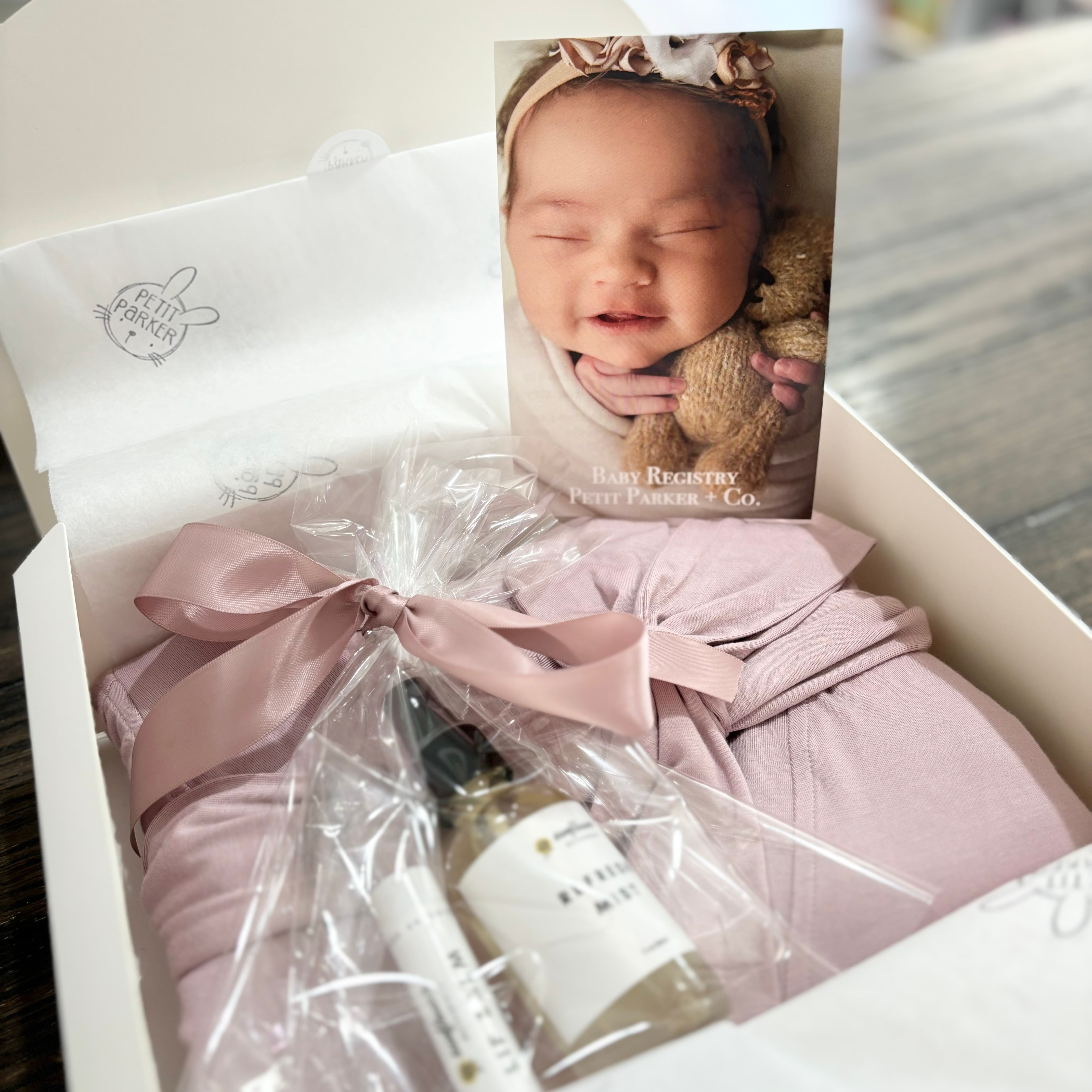 Baby in a box with a photo of a smiling baby girl on top, wrapped in pink cloth, with a small gift and a bottle inside.