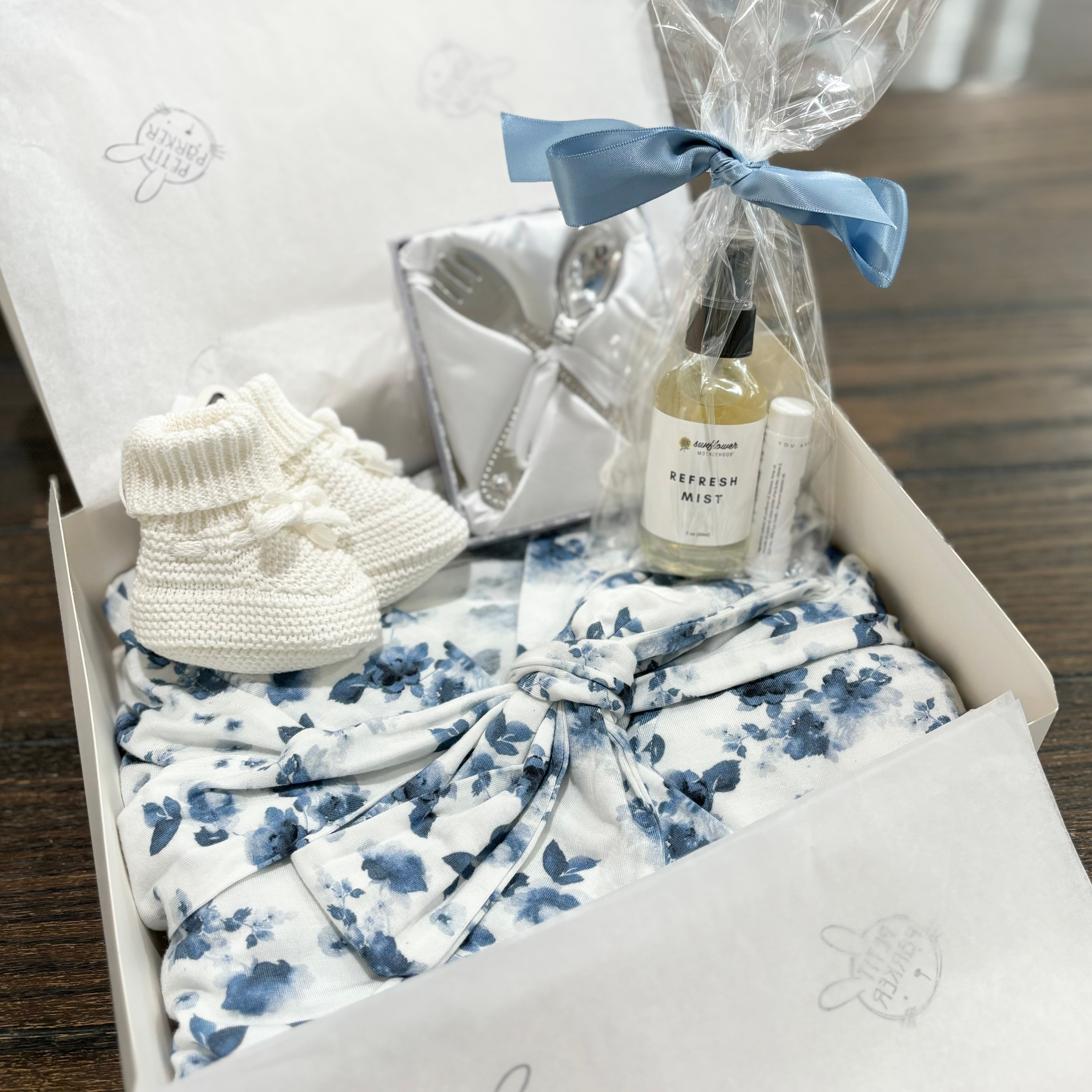 Gift box with baby shoes, floral clothing, a small bottle, and a wrapped gift with a blue ribbon.