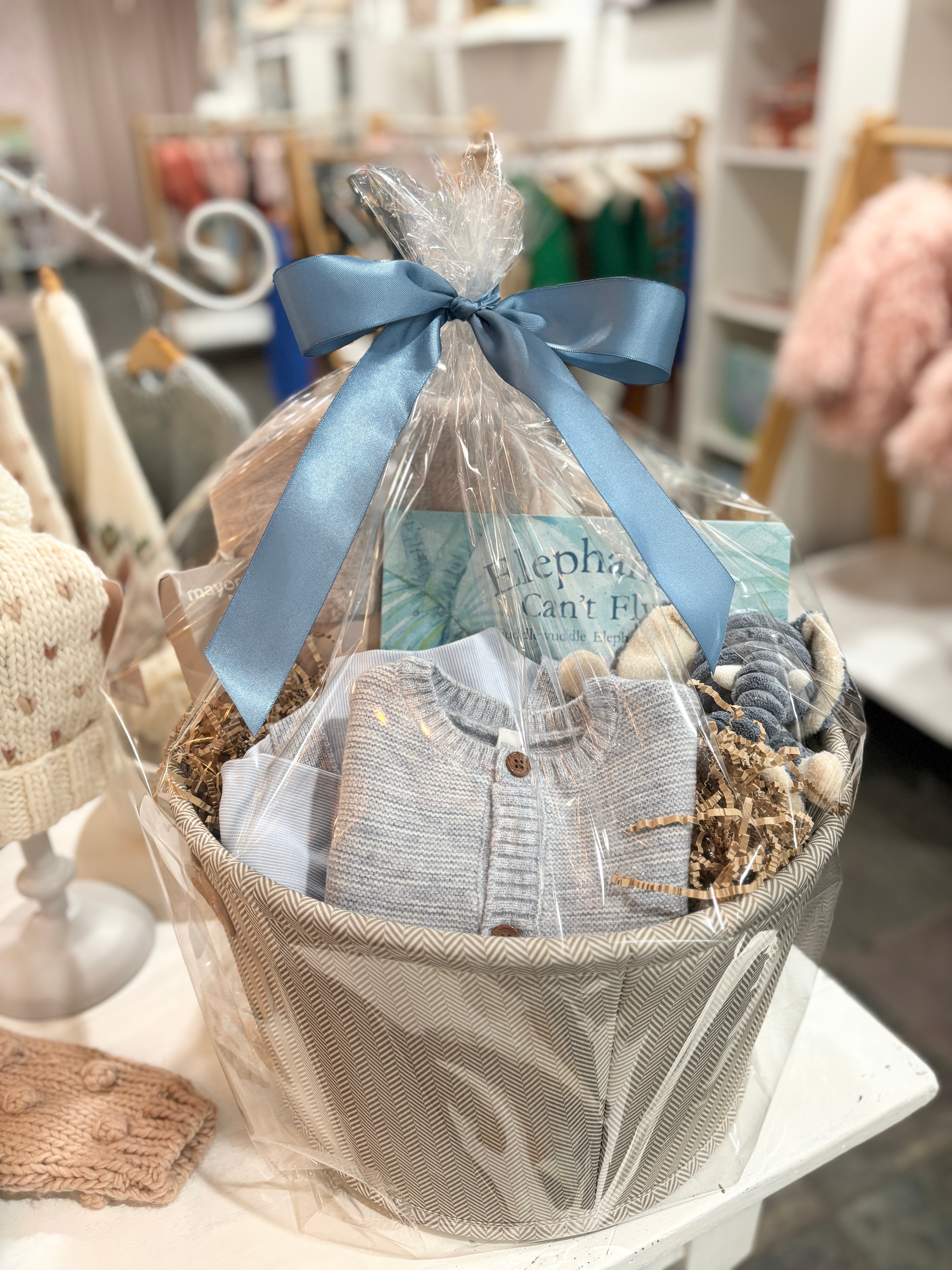 Gift basket with baby clothes and accessories, wrapped with a blue ribbon, displayed on a white surface in a store.