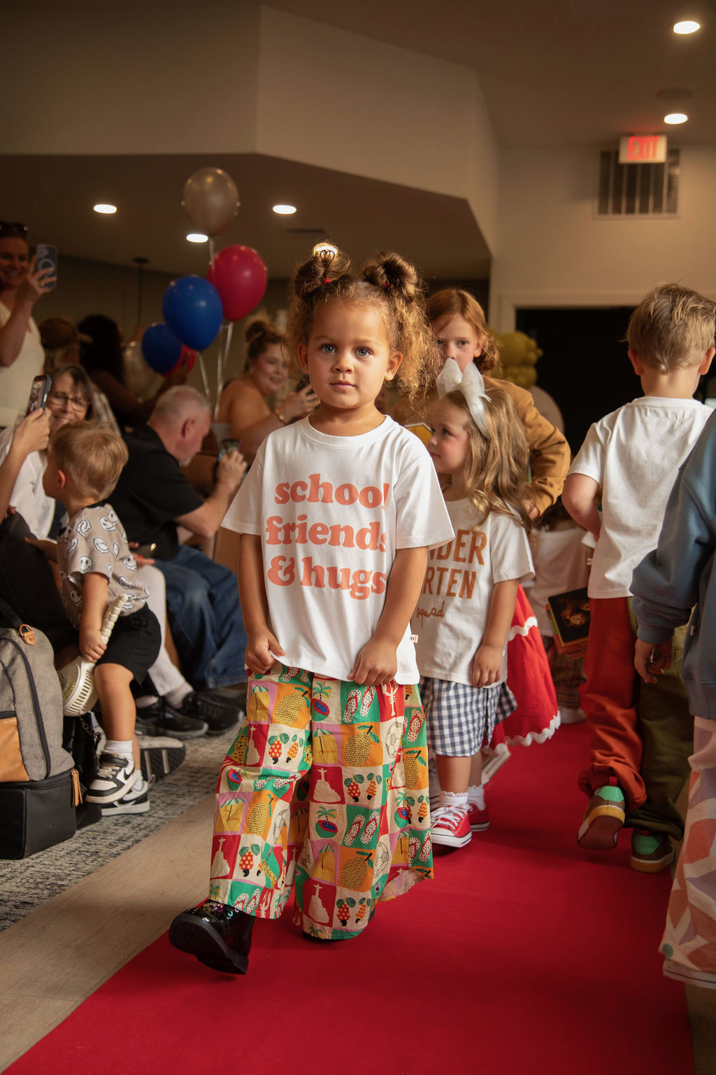 Young girl walking on a red carpet at a party with balloons and other children in the background.