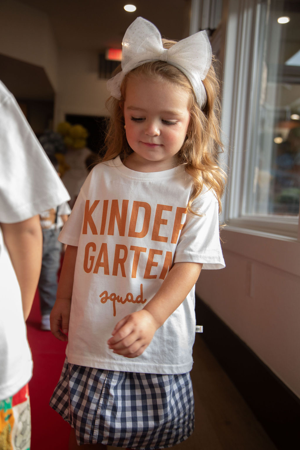 Young girl with a white bow in her hair, wearing a white T-shirt with orange text, standing indoors near a window.