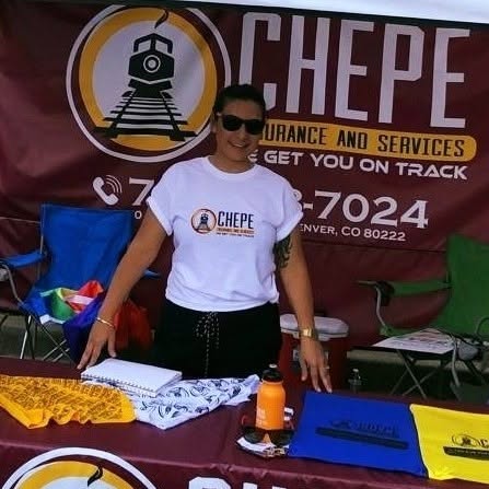 Woman standing behind a table with promotional materials, wearing sunglasses and a white T-shirt, at an outdoor event.