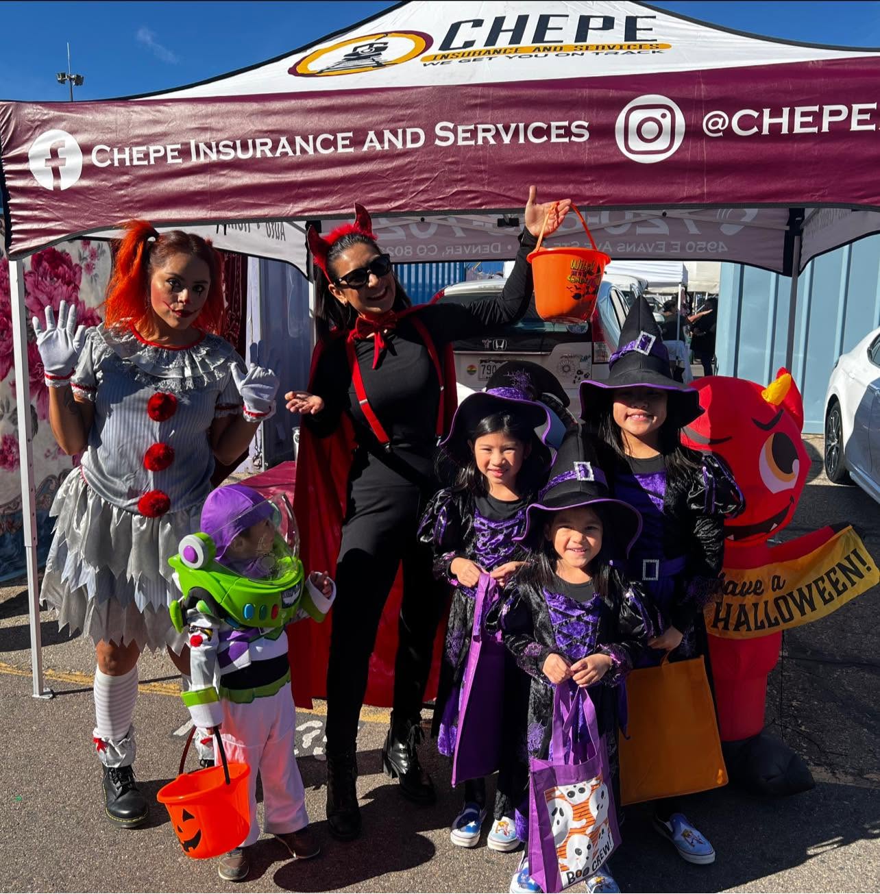 Group of people in Halloween costumes standing under a tent with CHEPE branding, holding buckets for trick-or-treating.
