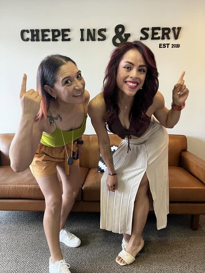 Two women smiling and pointing upward, standing in front of a wall with text, in a room with a brown sofa.