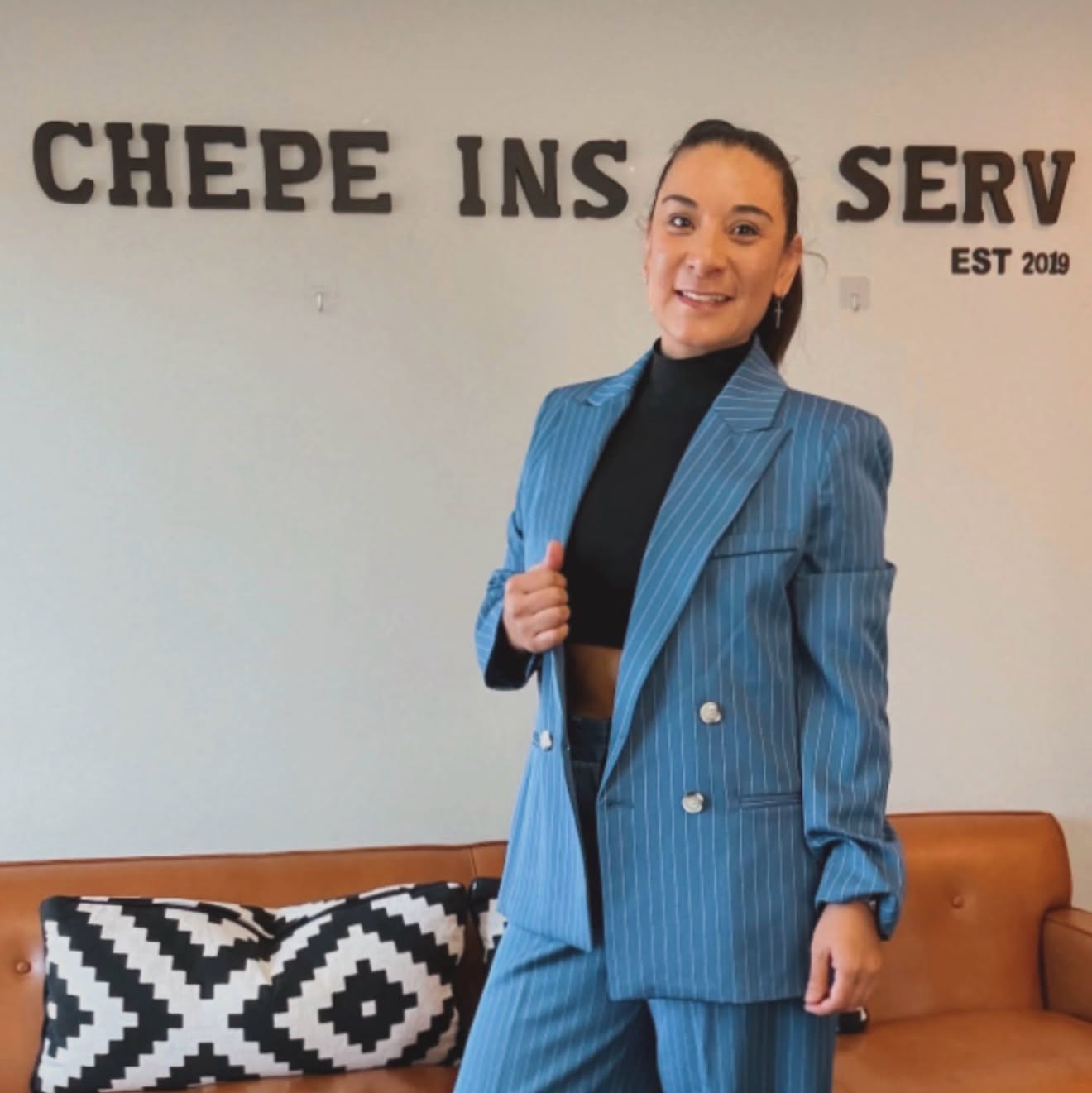 Woman in a blue pinstripe blazer and black turtleneck standing in front of a wall with partial text, smiling.