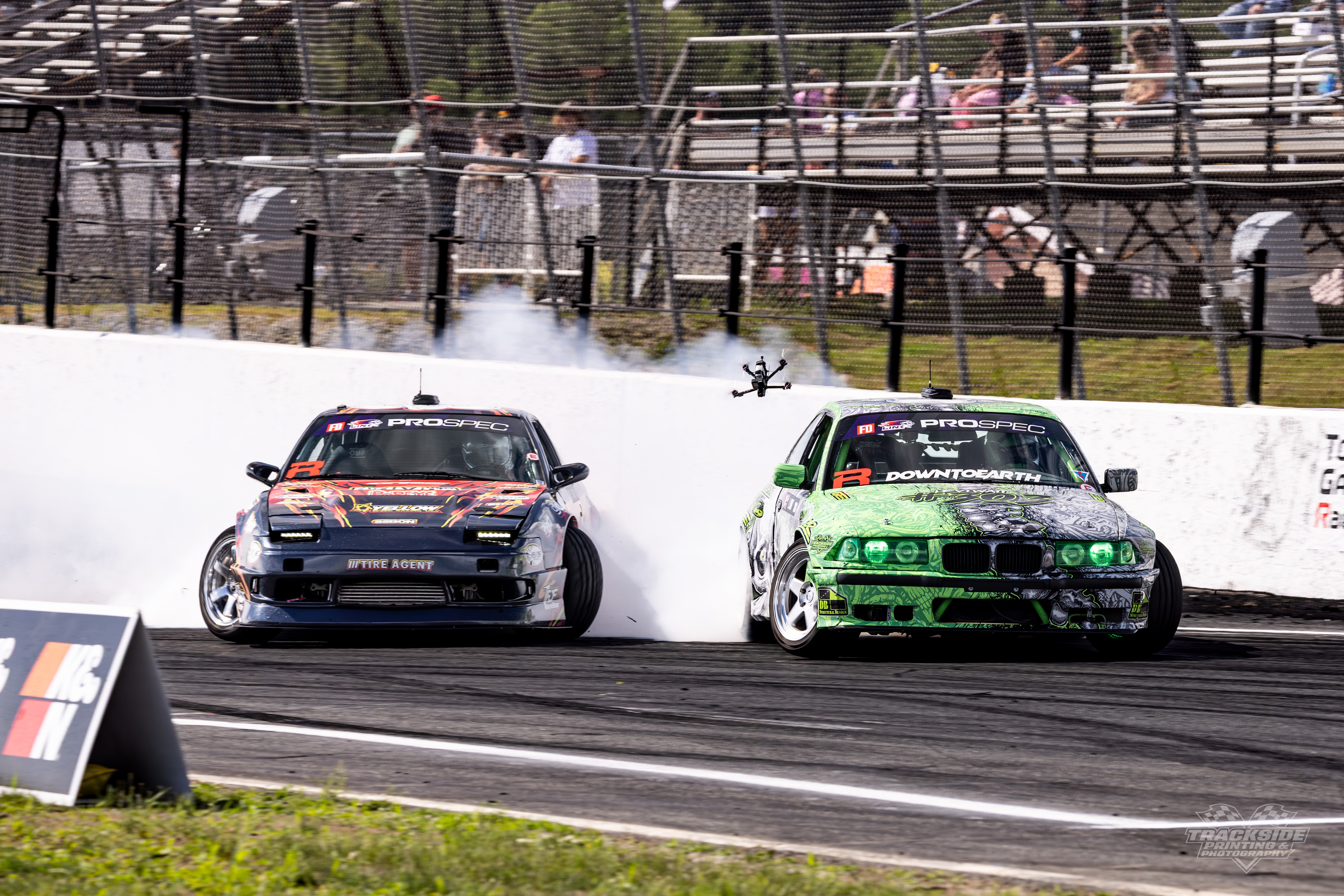 Two race cars collide on a track, with smoke and debris, while spectators watch from behind a fence.