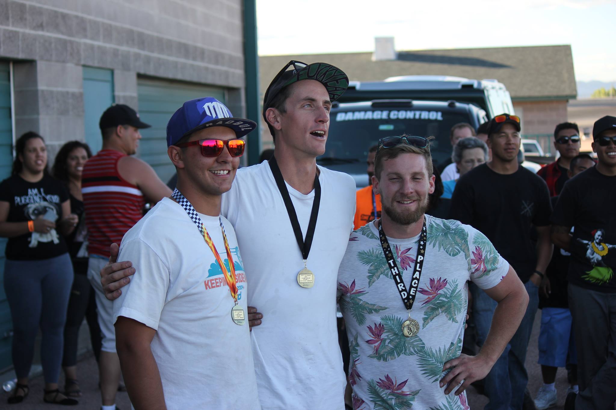 Three men with medals around their necks pose outdoors, with a group of people and a vehicle in the background.