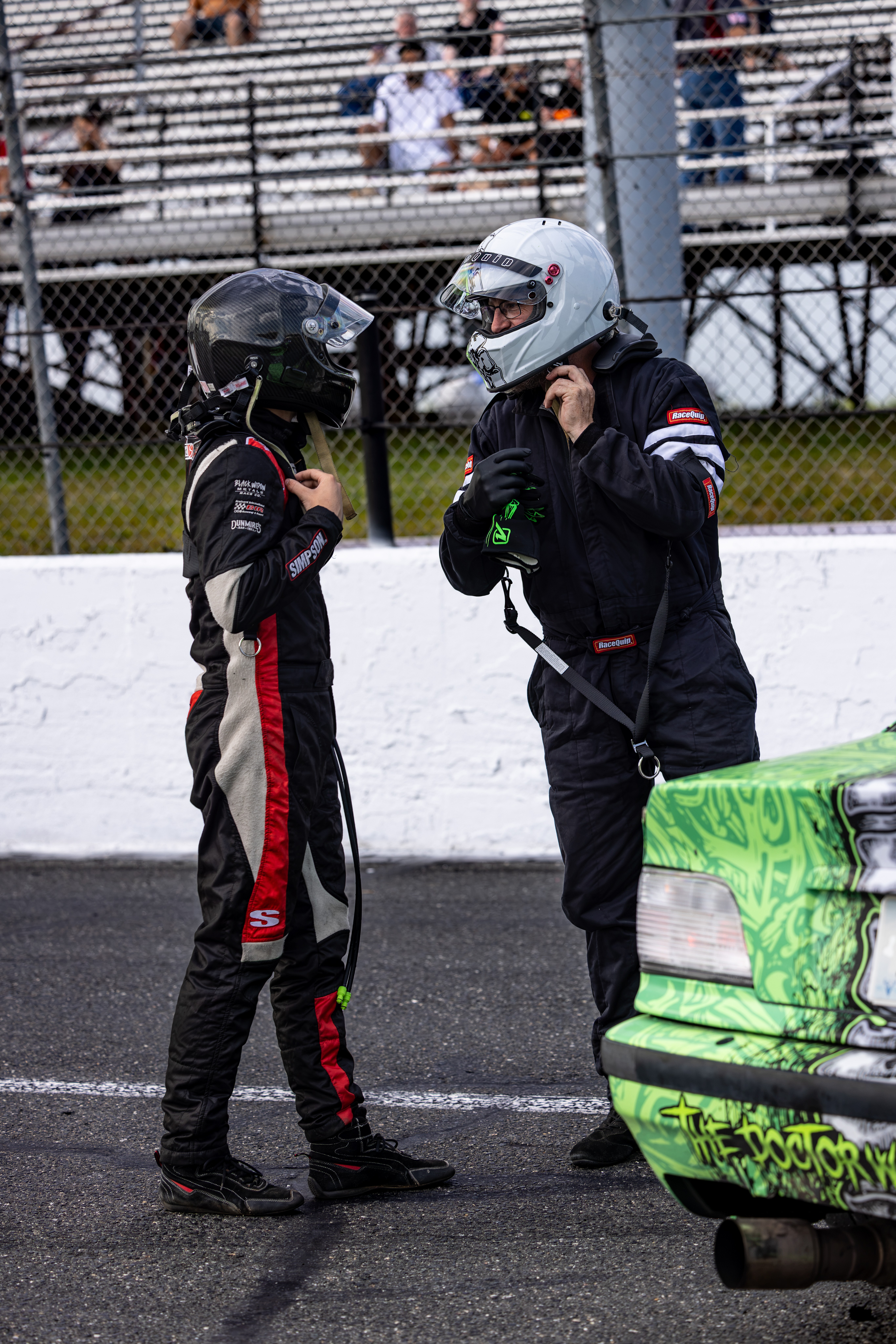Two race car drivers in racing suits and helmets stand on the track, talking near a green race car with Monster Energy branding.