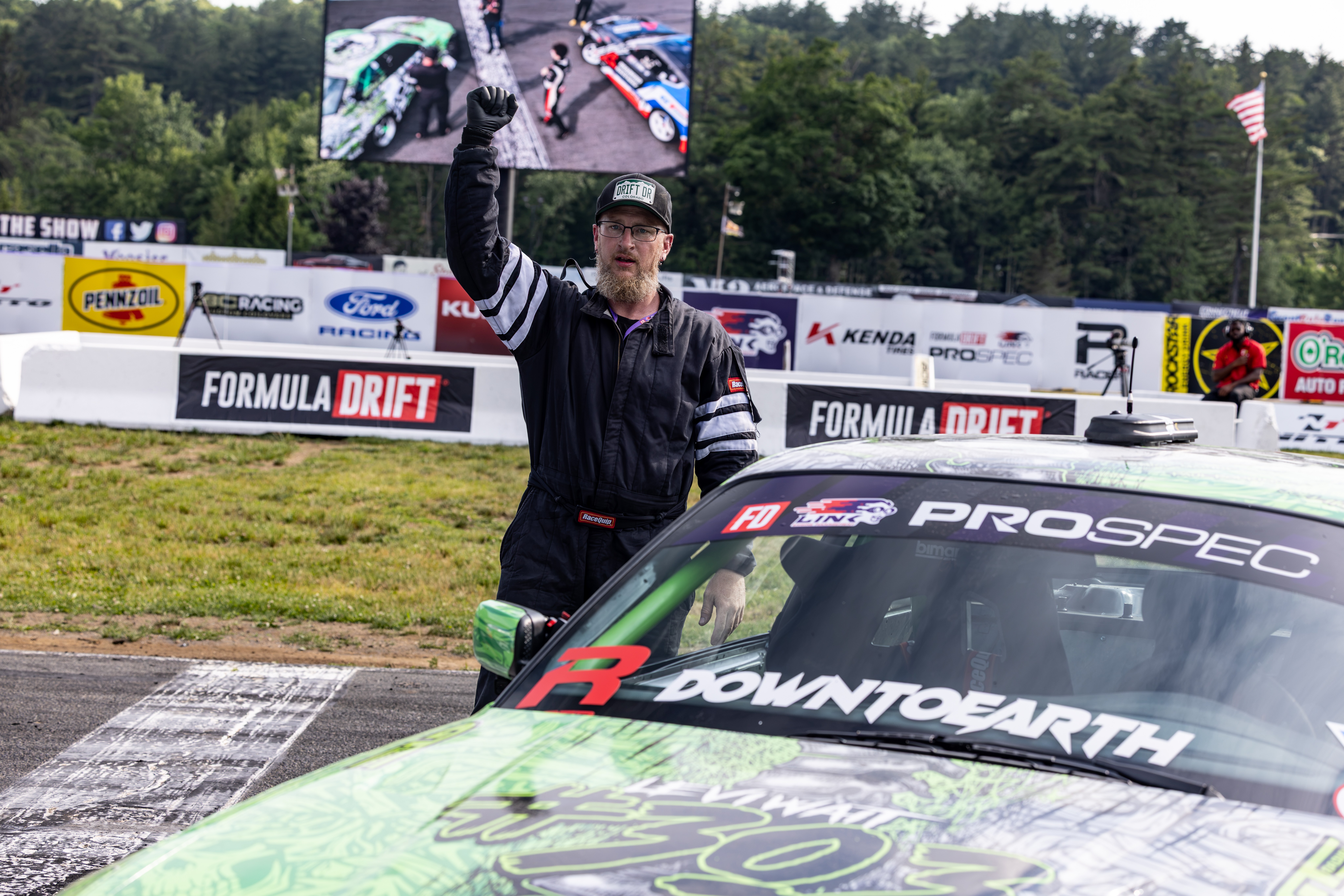 Race official holding a checkered flag at a racing track with a car in the foreground and a large screen above.