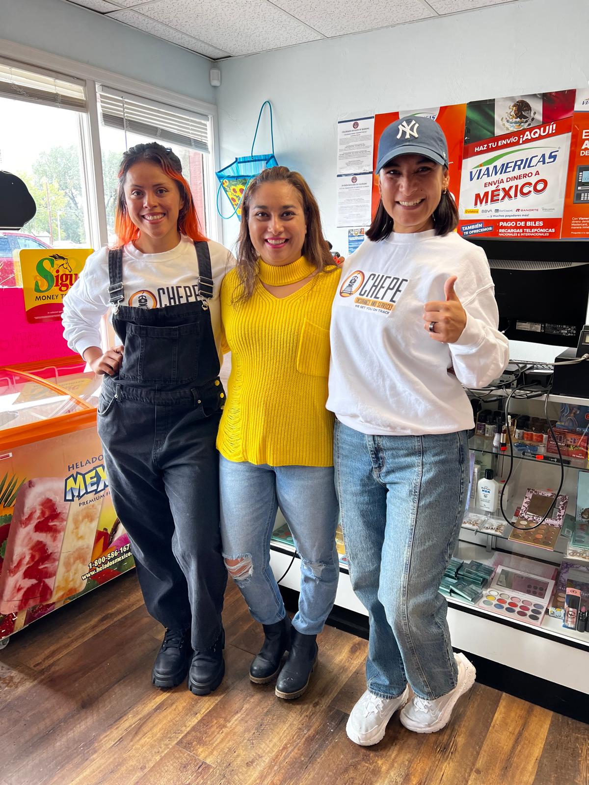 Three women standing indoors, smiling, with a window, posters, and a display case behind them.