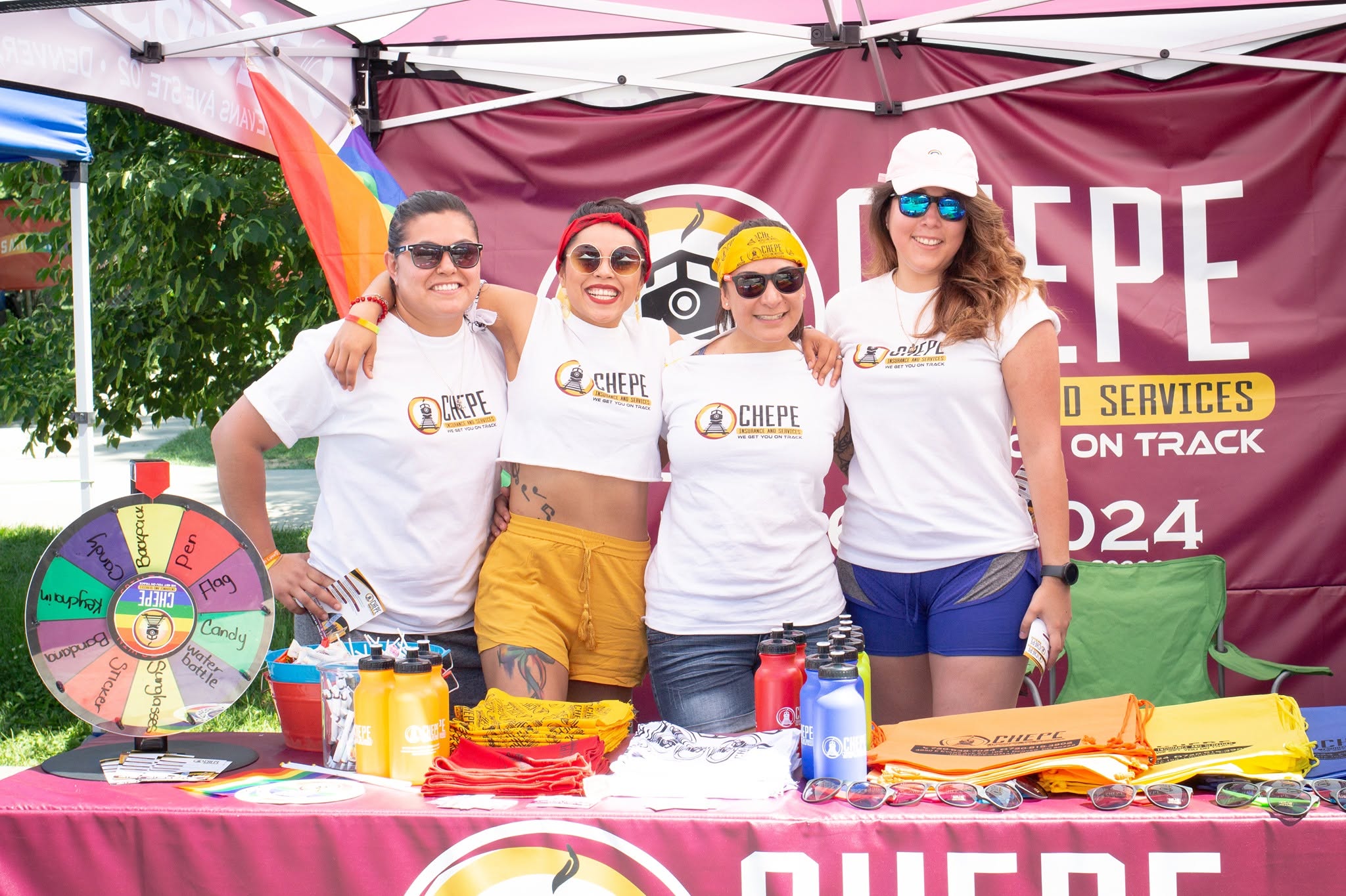 Four women wearing white shirts and colorful shorts stand behind a table at an outdoor event, smiling with arms around each other.