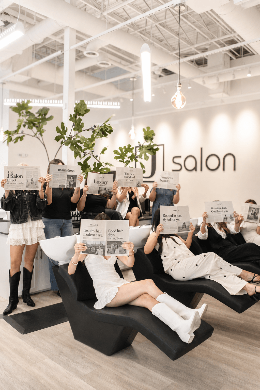 Group of children in salon holding newspapers, some sitting on black lounge chairs, with salon logo and plants in background.