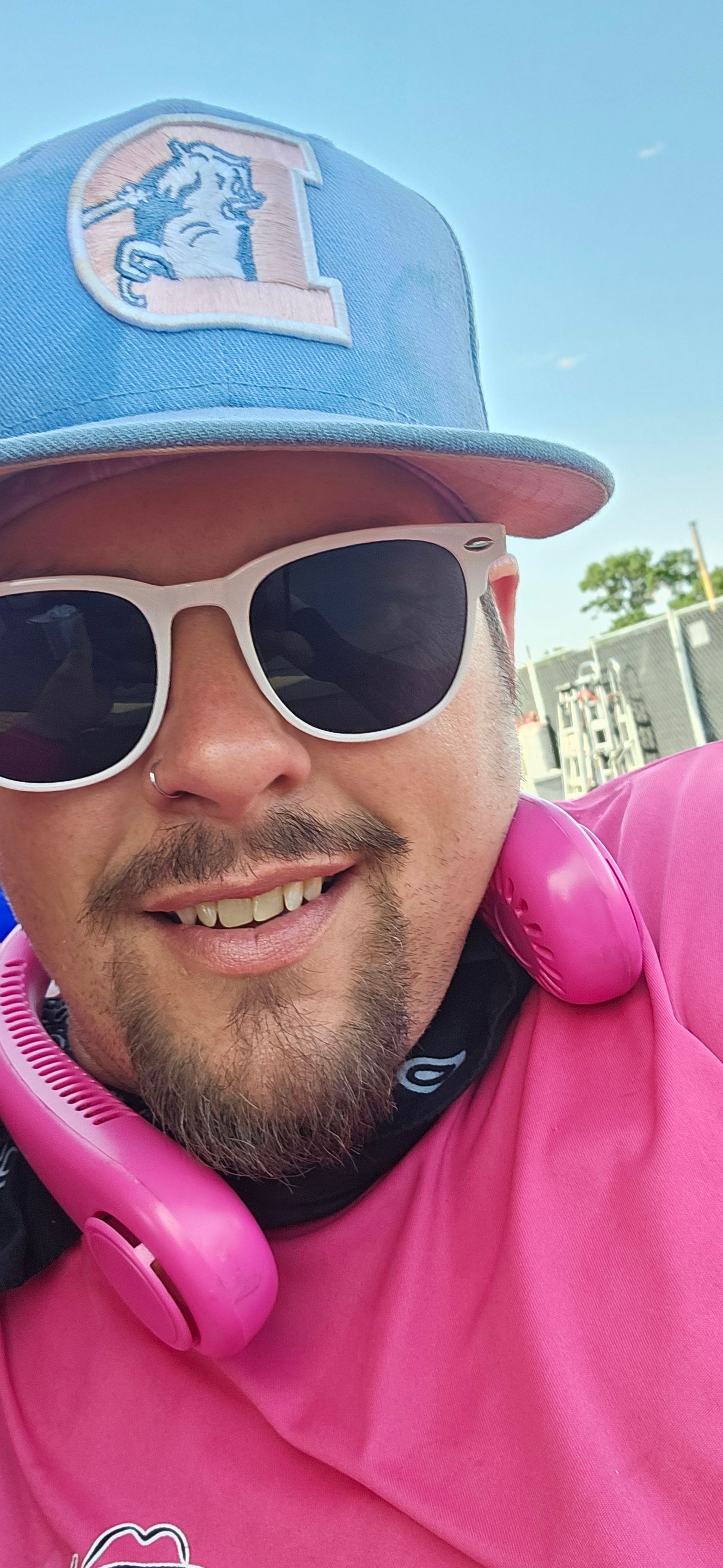 Person wearing sunglasses and a blue cap with a logo, smiling, with pink headphones, outdoors on a sunny day.