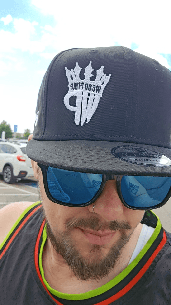 Person wearing a black cap with a white logo, sunglasses, and a colorful shirt, outdoors with cars in background.