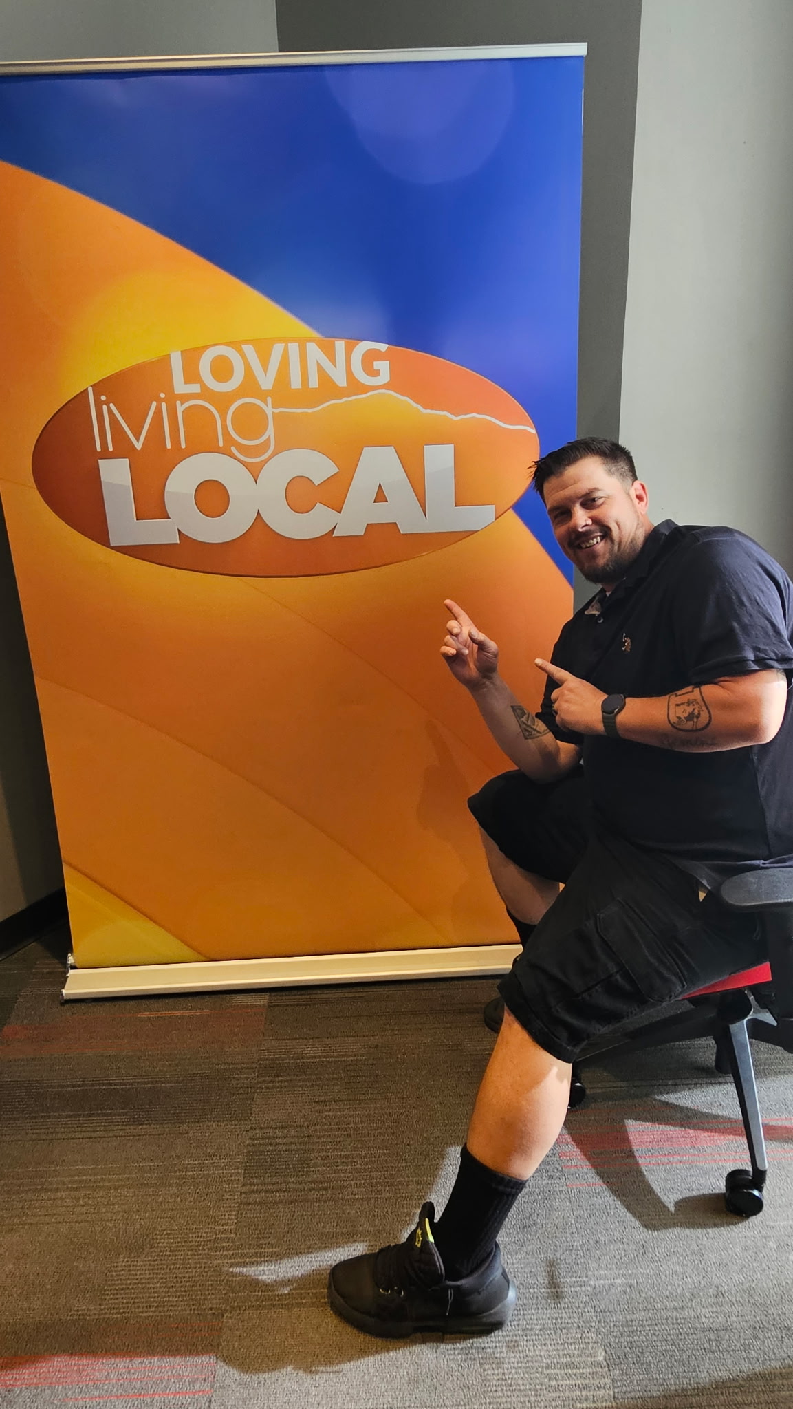 Man sitting on a stool next to a colorful sign with the text 'Loving Living Local'.