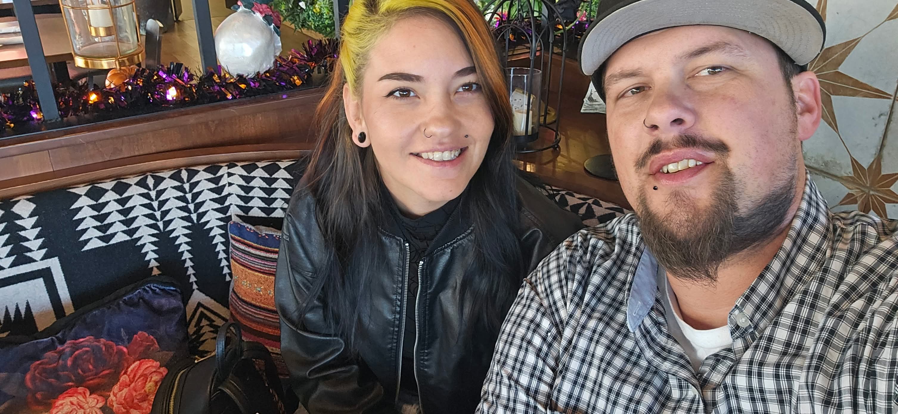 Two people smiling, sitting indoors with colorful decorations and patterned cushions behind them.