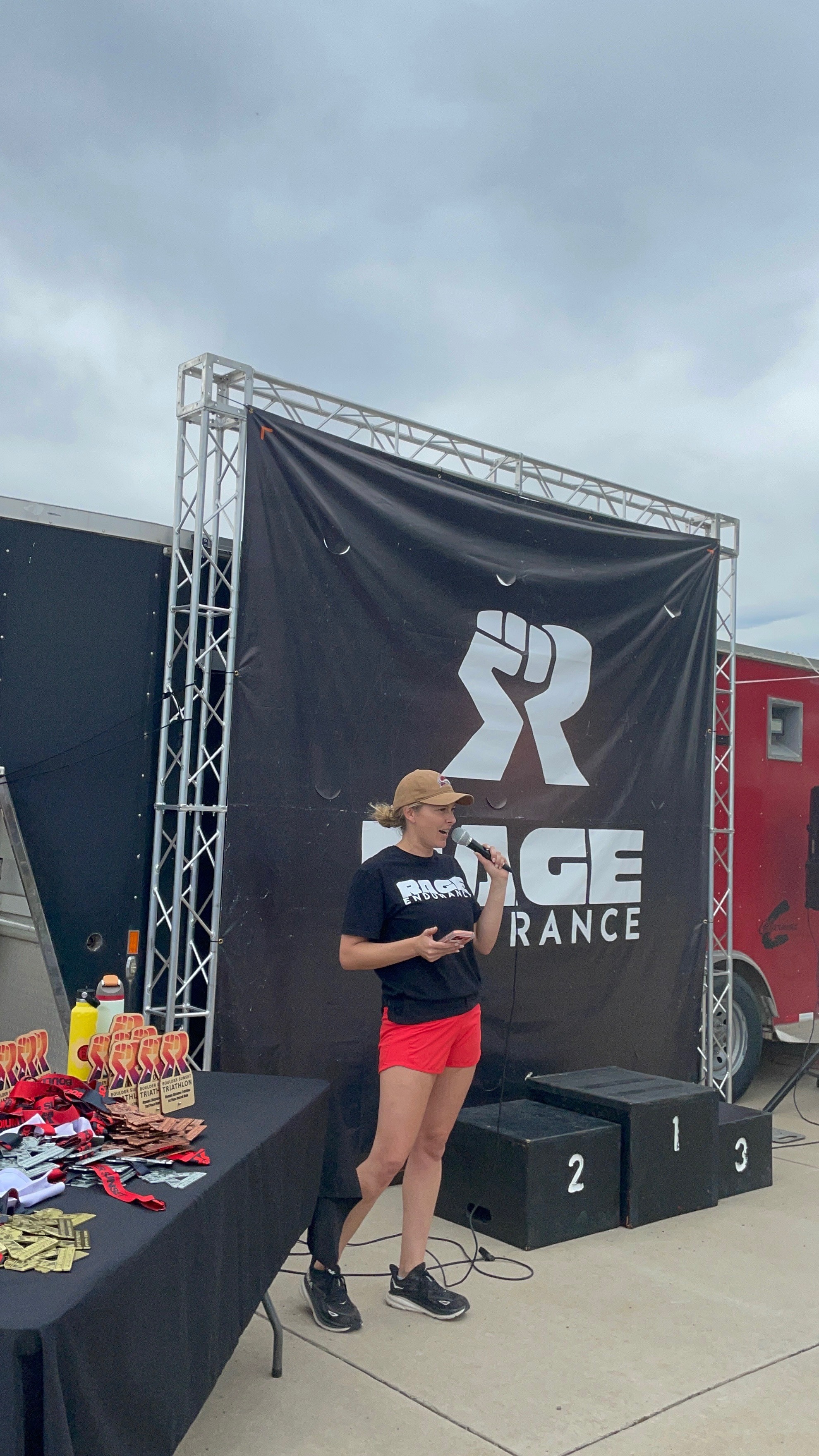 Person standing on a stage with a large black banner, wearing a black shirt and red shorts, in an outdoor setting.