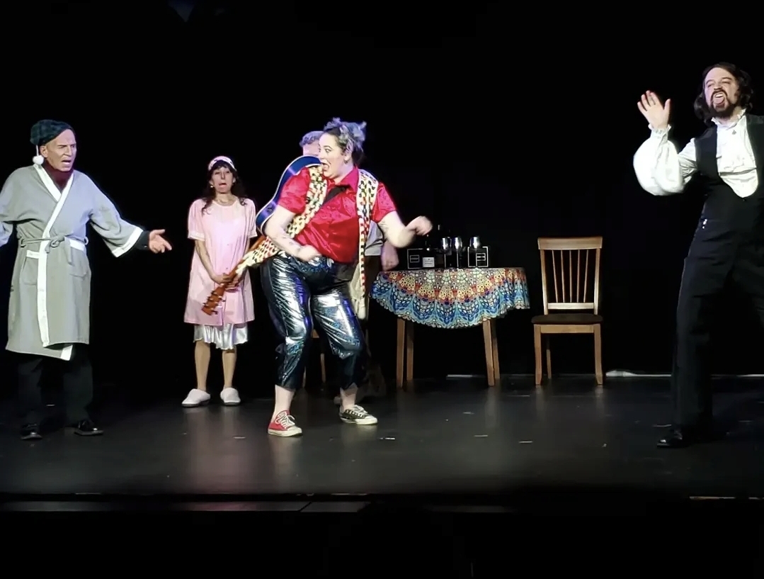 Four performers on stage, one playing a flute, others dancing and singing, with a table and chairs in the background.