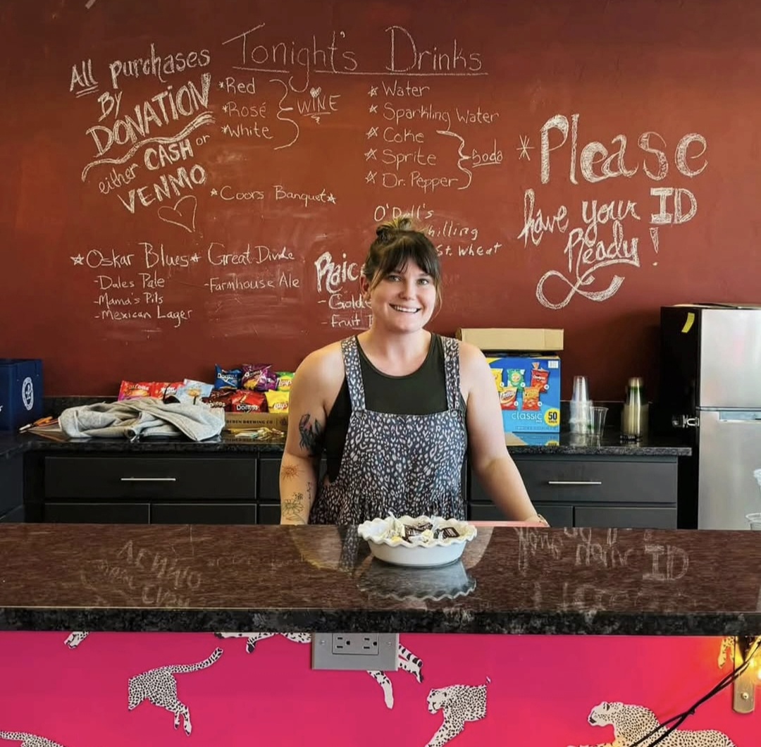 Young woman standing behind a counter in a cafe, smiling, with a red chalkboard menu behind her.