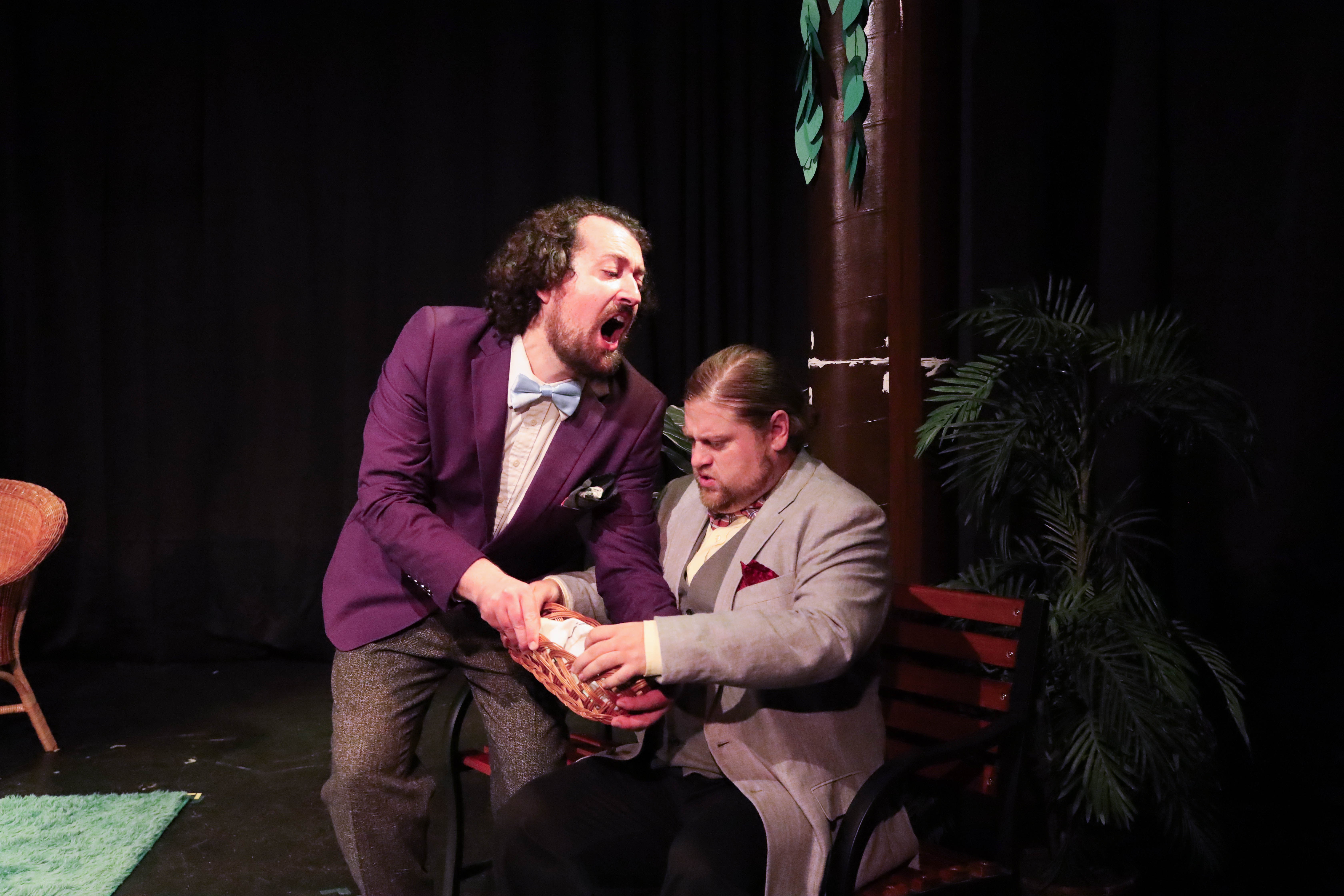 Two men on stage, one standing and shouting, the other sitting, holding a glass, with dark background and plants.
