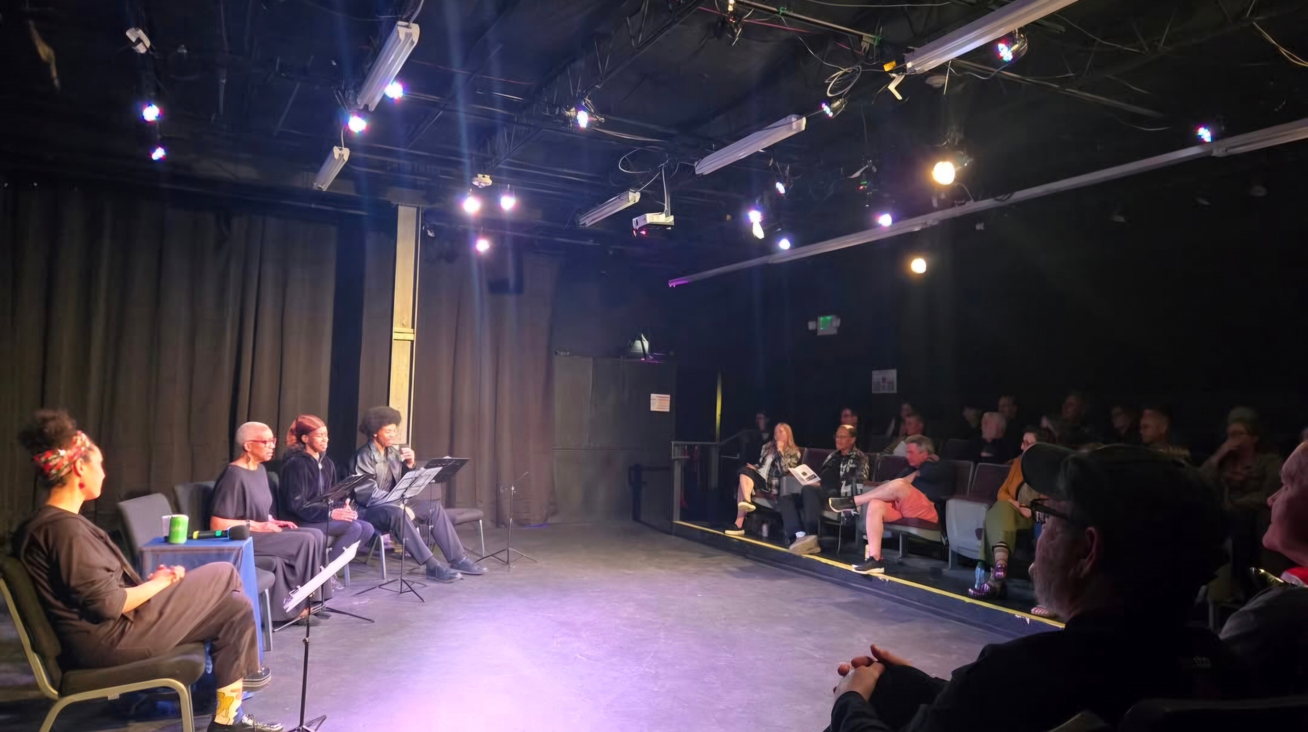 People seated on stage and in audience in a dark theater with stage lights, black curtains, and a black floor.
