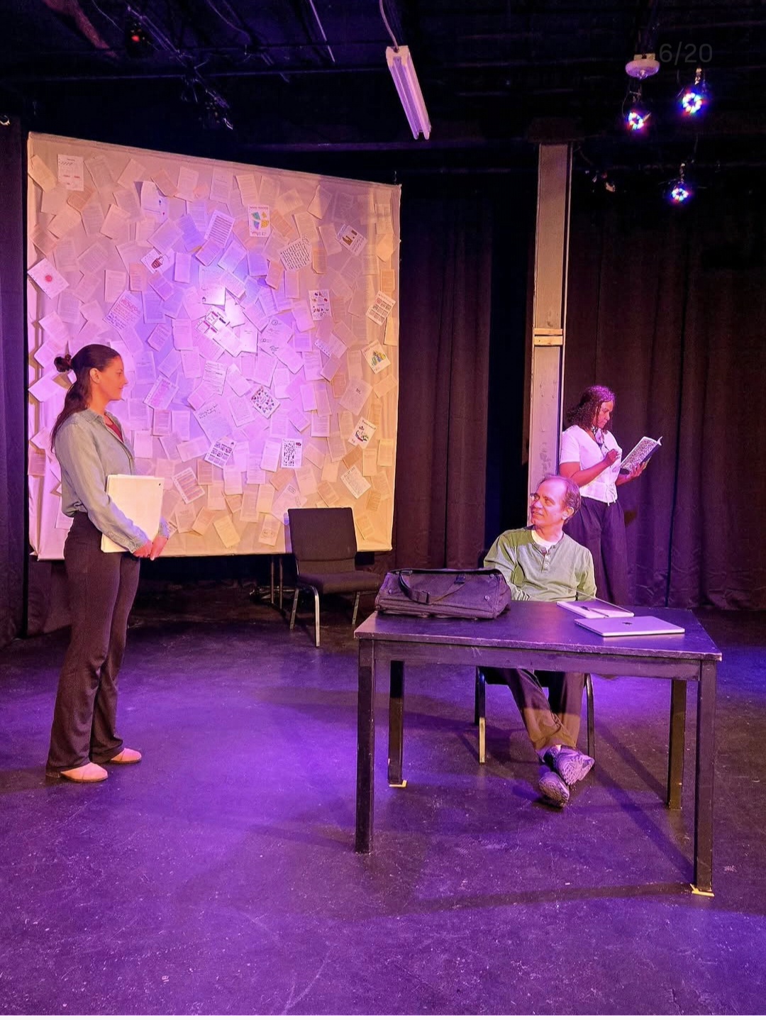 Three people on stage with a large screen, one sitting at a table, two standing, one reading from a paper.