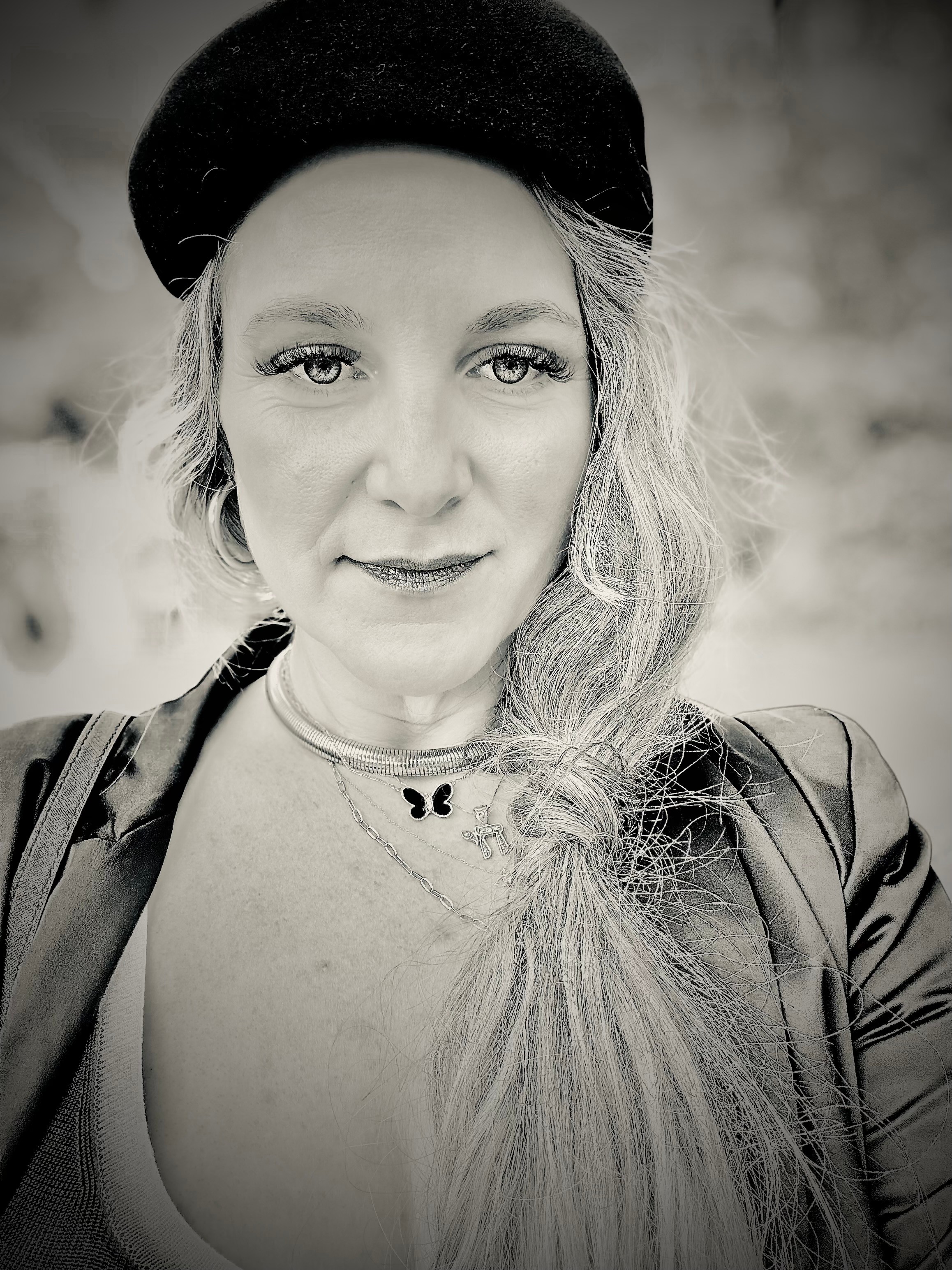 Woman with long hair wearing a beret and leather jacket, looking at camera, outdoors background, black and white.