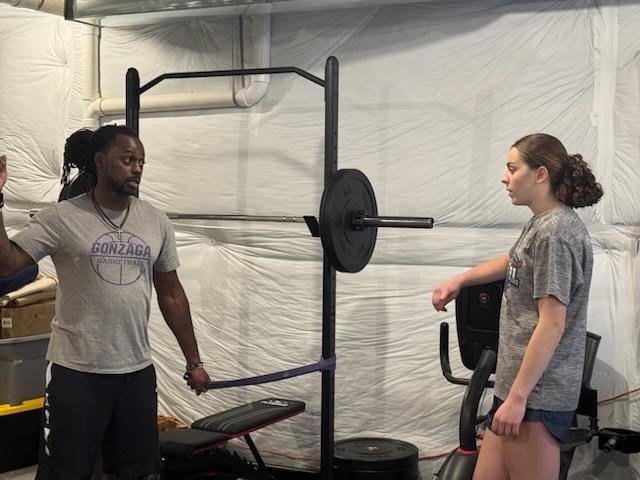 Two women in a gym with weightlifting equipment, one standing and the other sitting on a bench, facing each other.