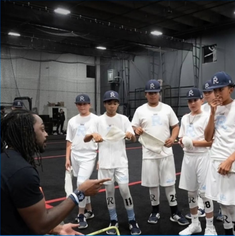 Group of young baseball players in white uniforms and blue caps listening to coach in indoor facility.