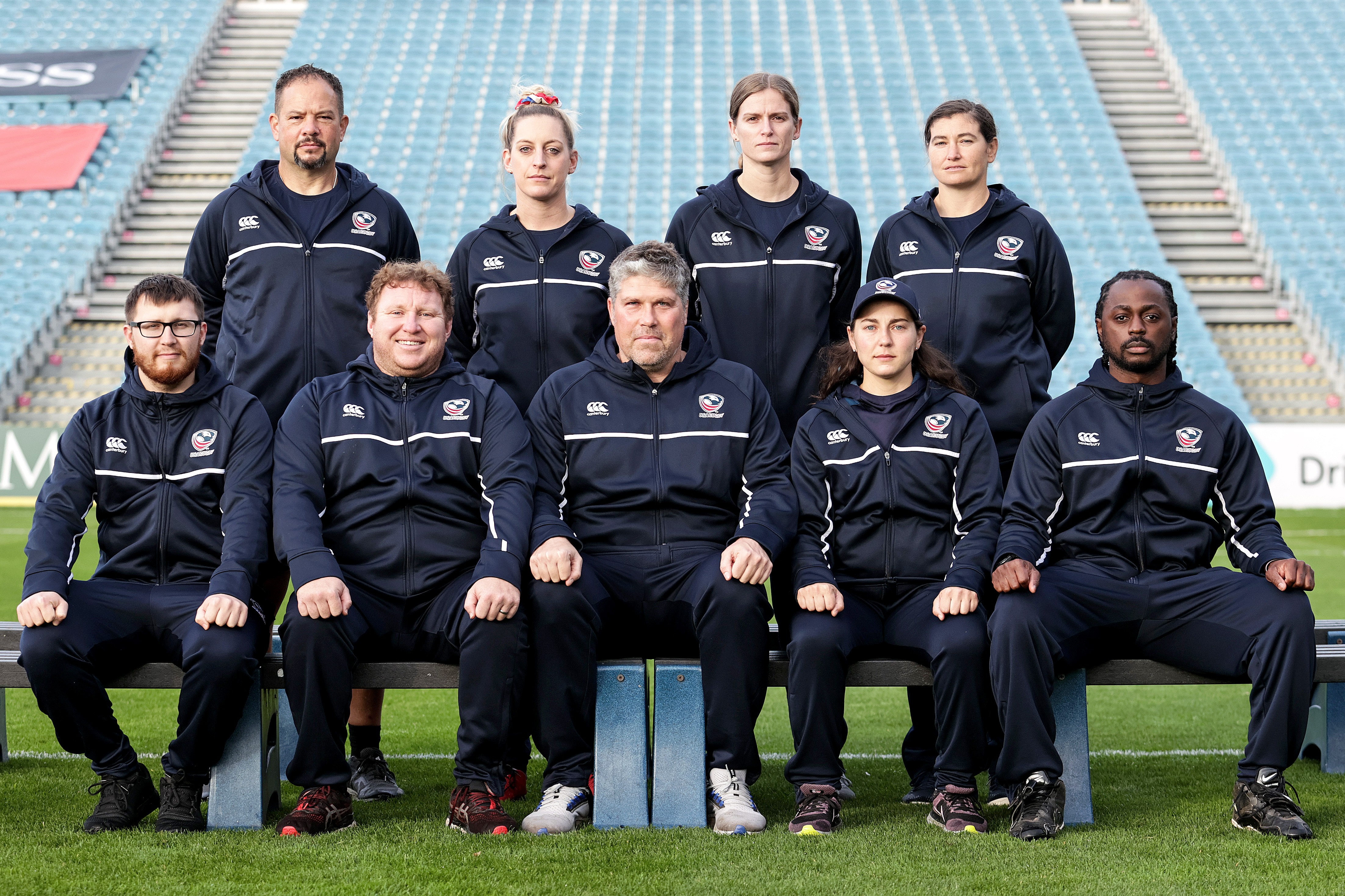 Group of nine people in matching dark sports jackets, seated and standing on a sports field with stadium seats in background.