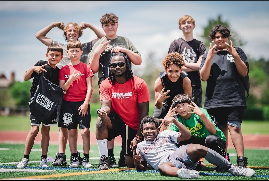 Group of children and a coach posing outdoors on a sports field, some making hand gestures, smiling.