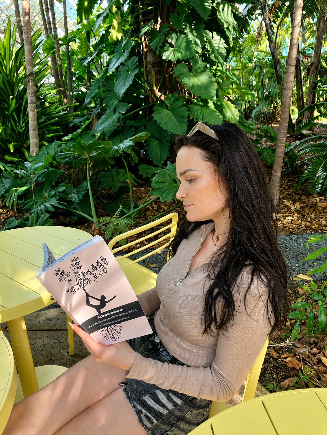 Young woman with long dark hair reading a book outdoors surrounded by lush green plants.