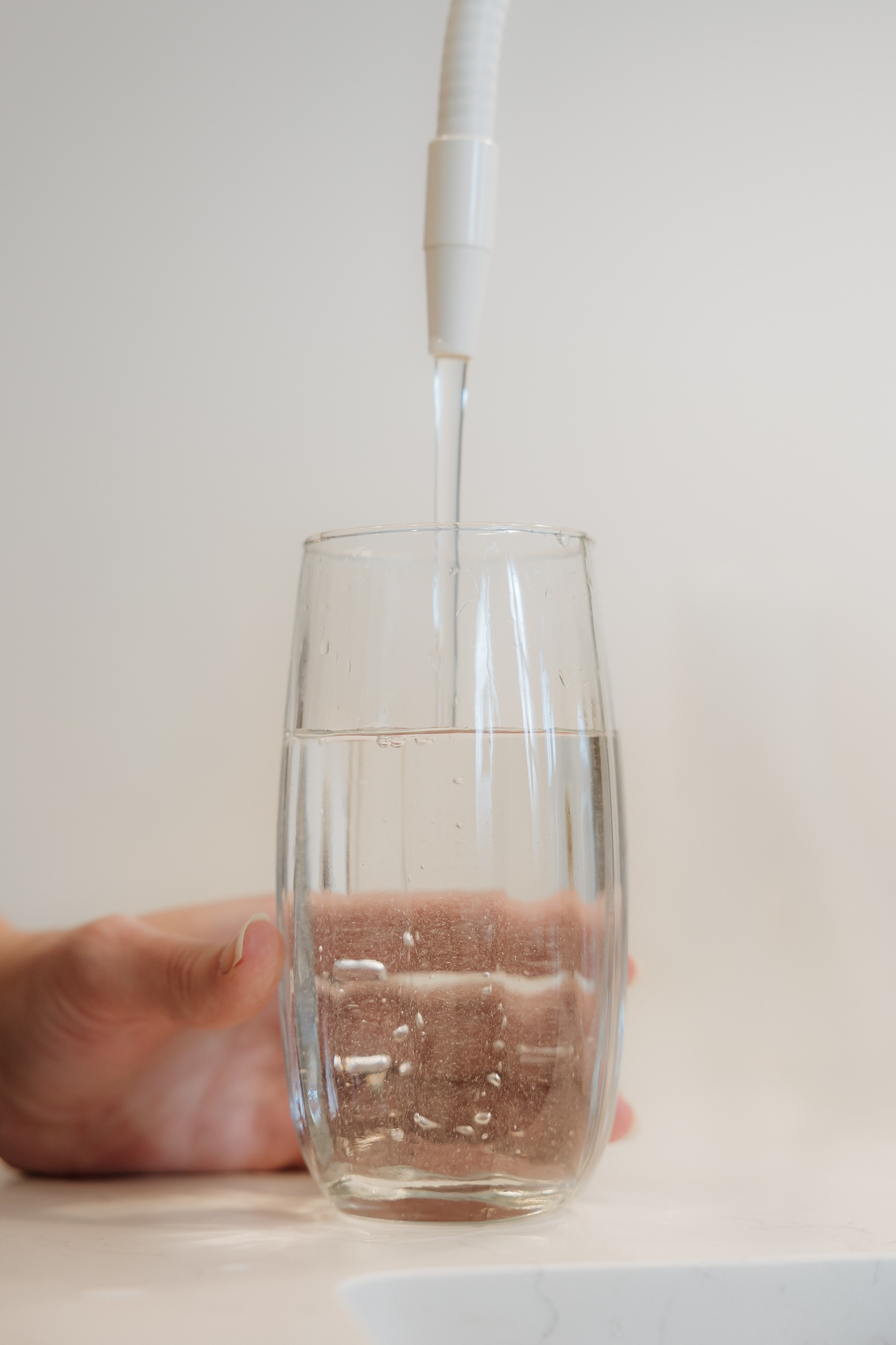 Hand holding a glass under a water faucet with water flowing into it, against a plain background.