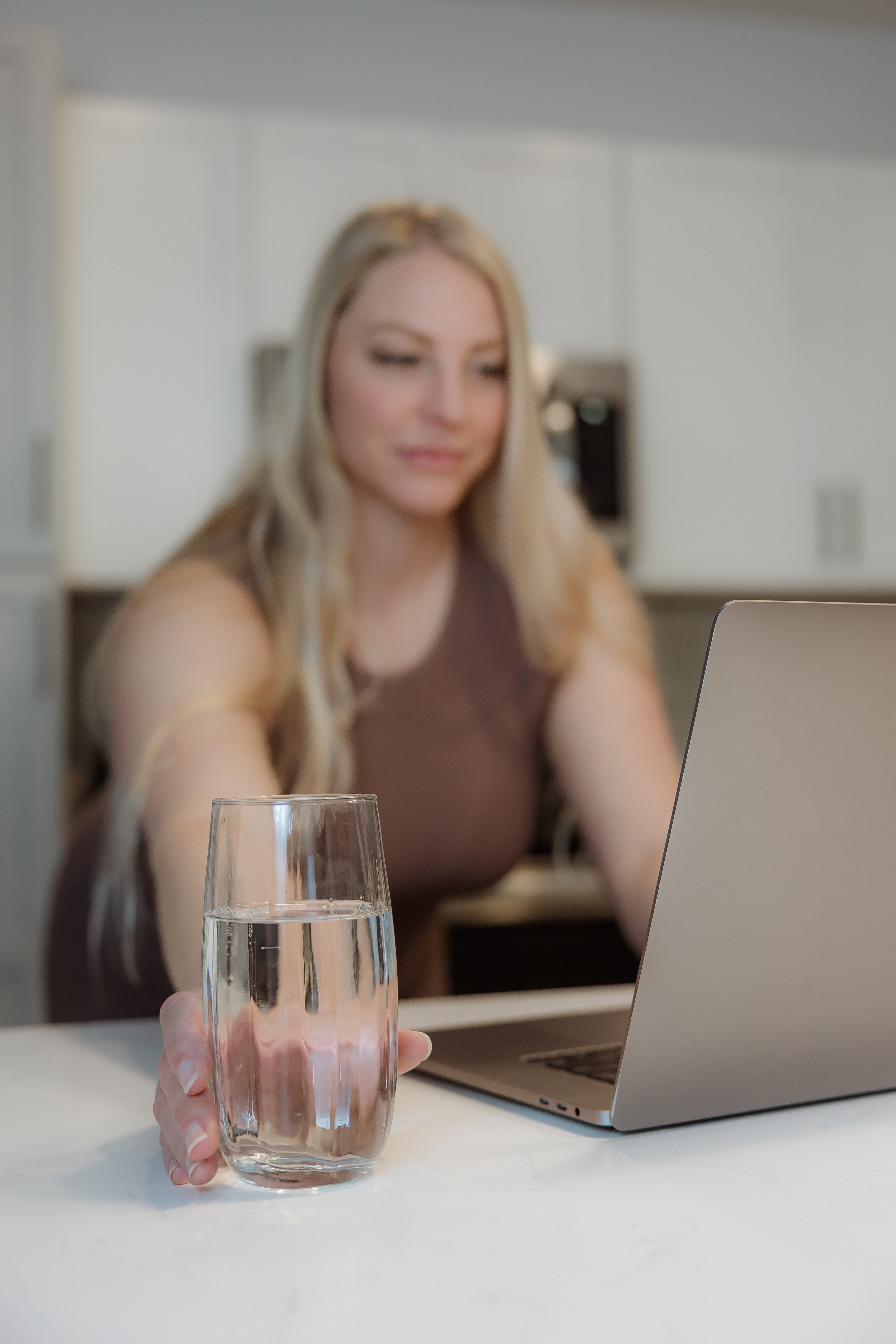 Woman with blonde hair working on a laptop at a kitchen counter, with a glass of water in front.