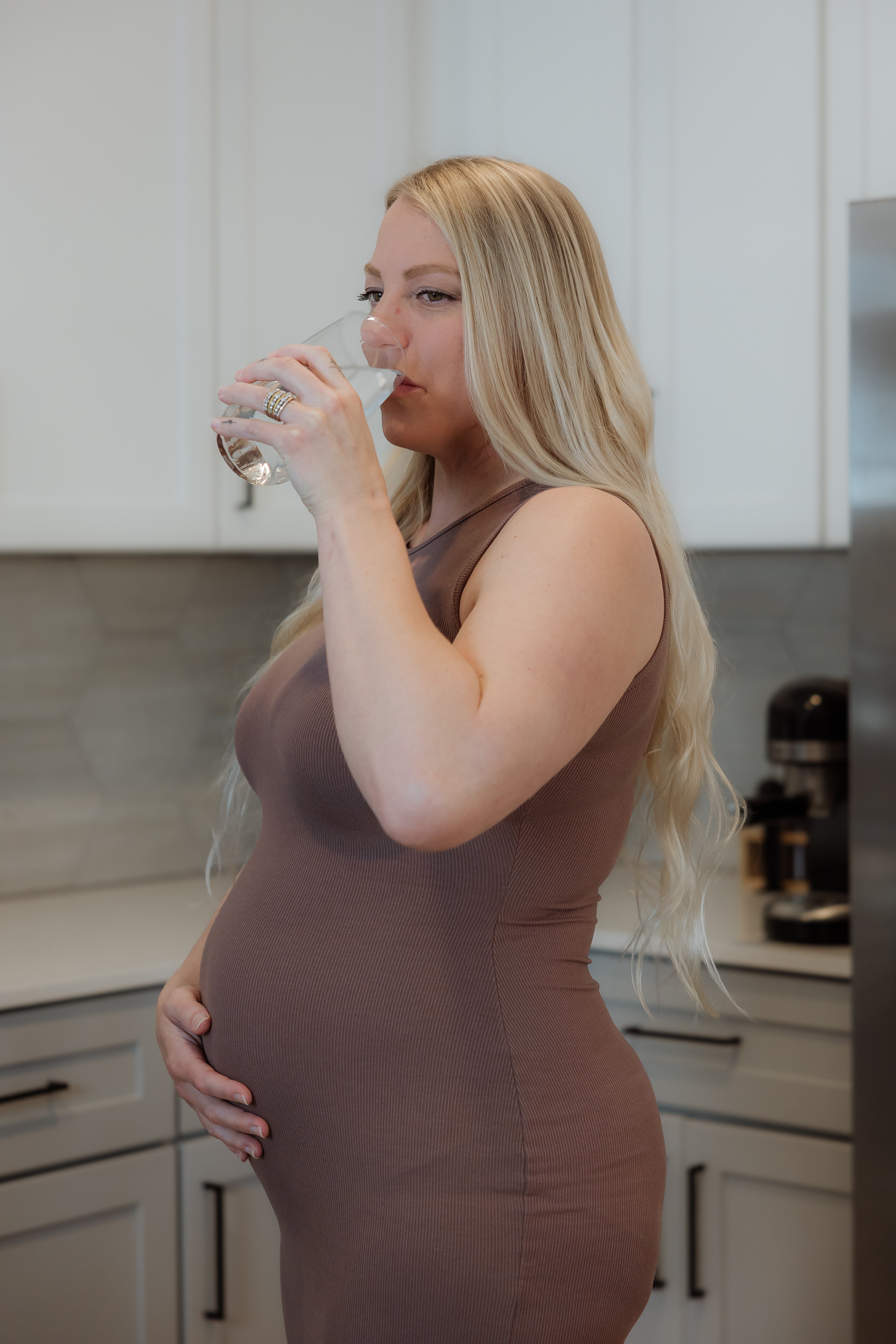 Woman with long blonde hair drinking from a glass in a kitchen, holding her stomach.